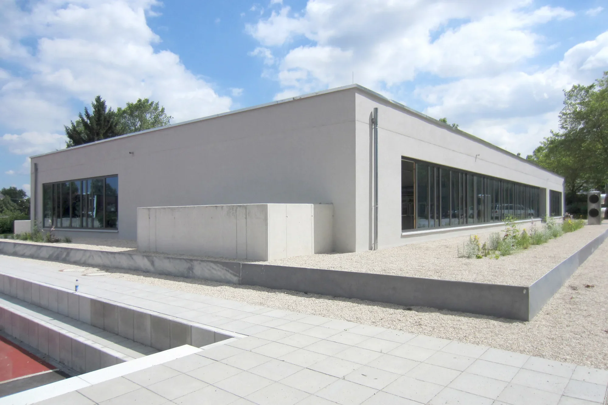 Ein modernes, weißes Gebäude mit großen Fensterfronten, umgeben von einem gepflasterten Vorplatz und einigen kleinen Büschen, bei sonnigem Himmel mit Wolken.