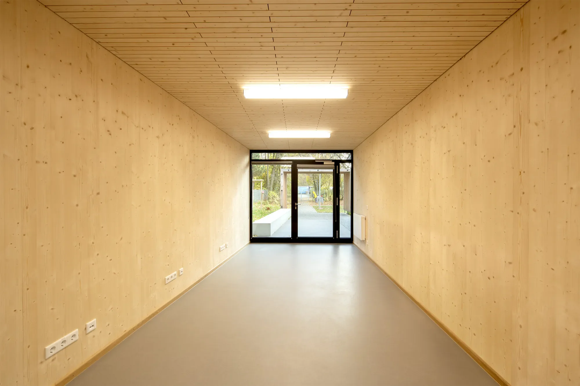Leeres Tunnel mit Holzdecke und Wänden, Beleuchtung, Glas-Tür am Ende mit Blick auf einen Gehweg und Bäume im Freien.