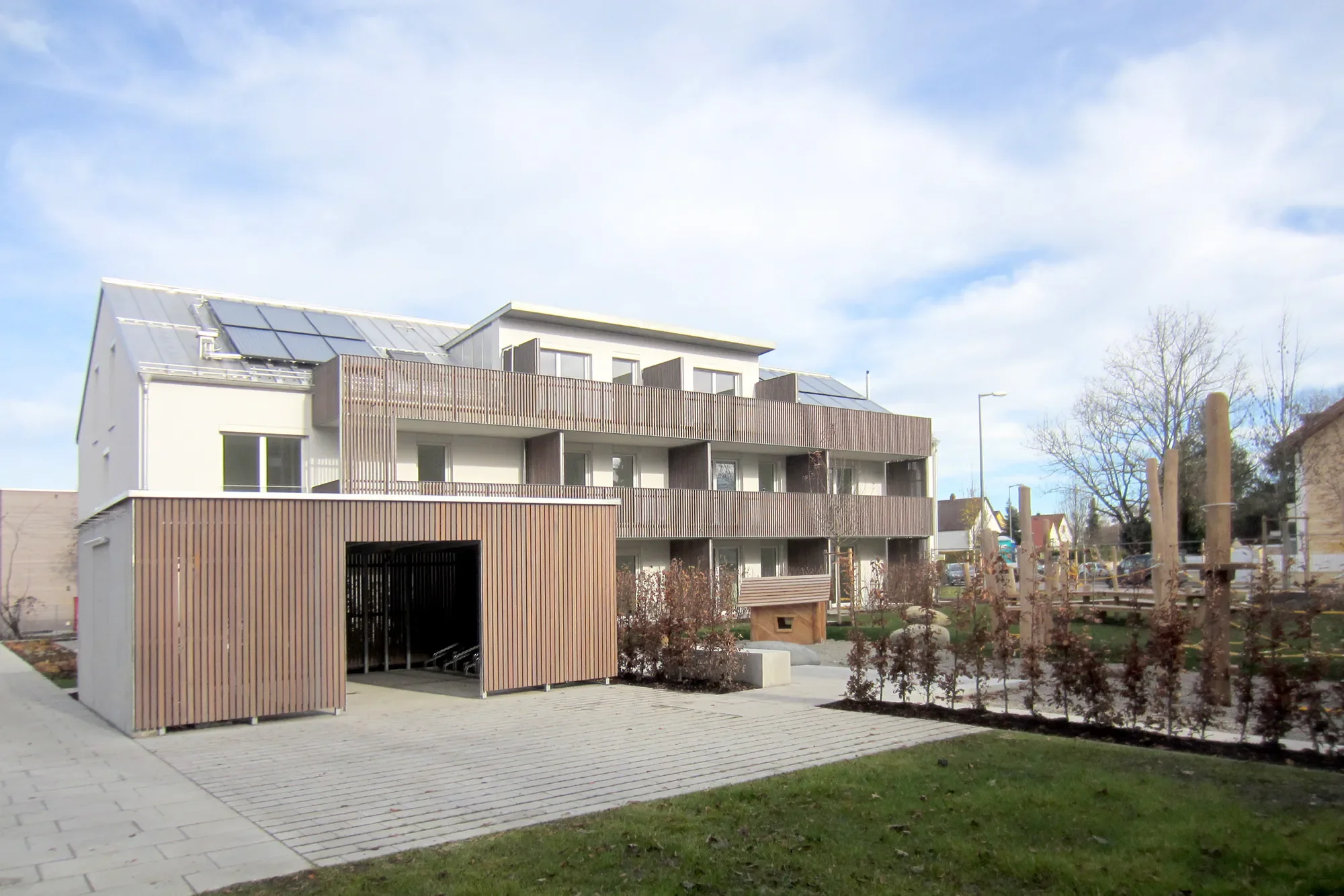 Moderner vierstöckiger Wohnblock mit Holzverkleidungen und Solarzellen auf dem Dach, umgeben von einem Garten mit Bäumen und einem gepflasterten Bereich