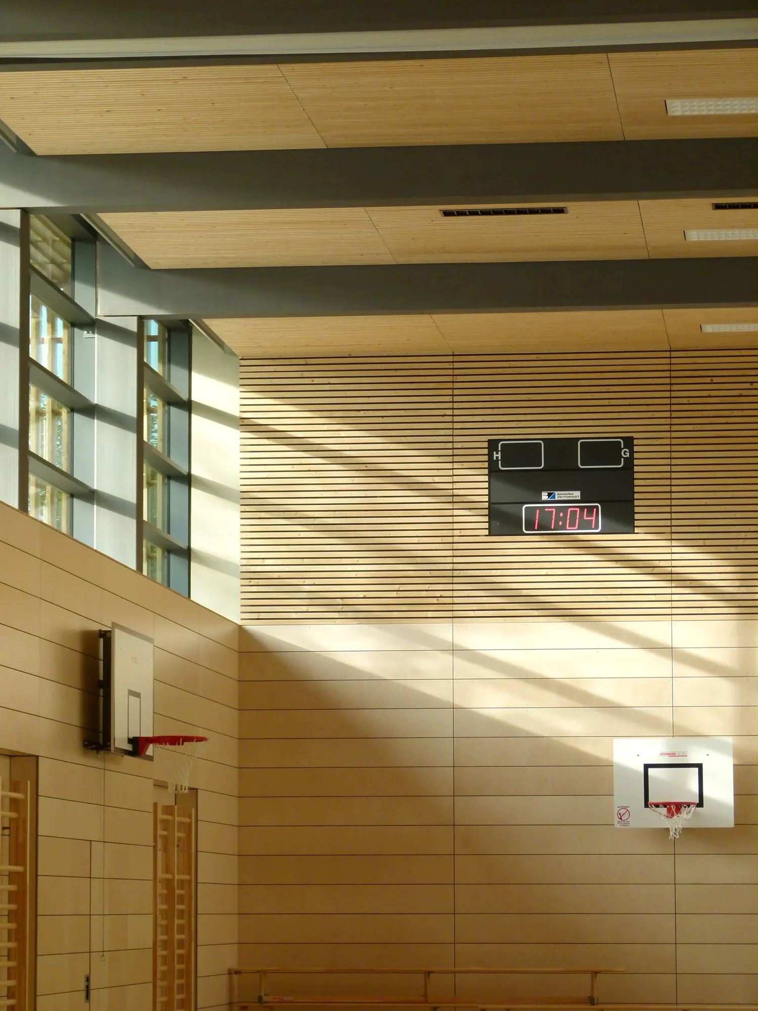 Innenraum eines Sportzentrums mit Holzwänden, Basketballkörben, einer Anzeigetafel mit der Uhrzeit 17:04, und natürlichen Licht durch große Fenster