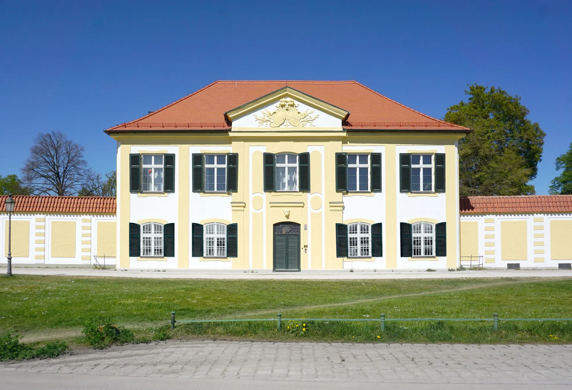 Ein barockes Haus mit gelber Fassade, weißen Fensterrahmen und schwarzen Fensterläden, rotes Ziegeldach, umgeben von Bäumen und einem grünen Rasen, bei blauem Himmel.