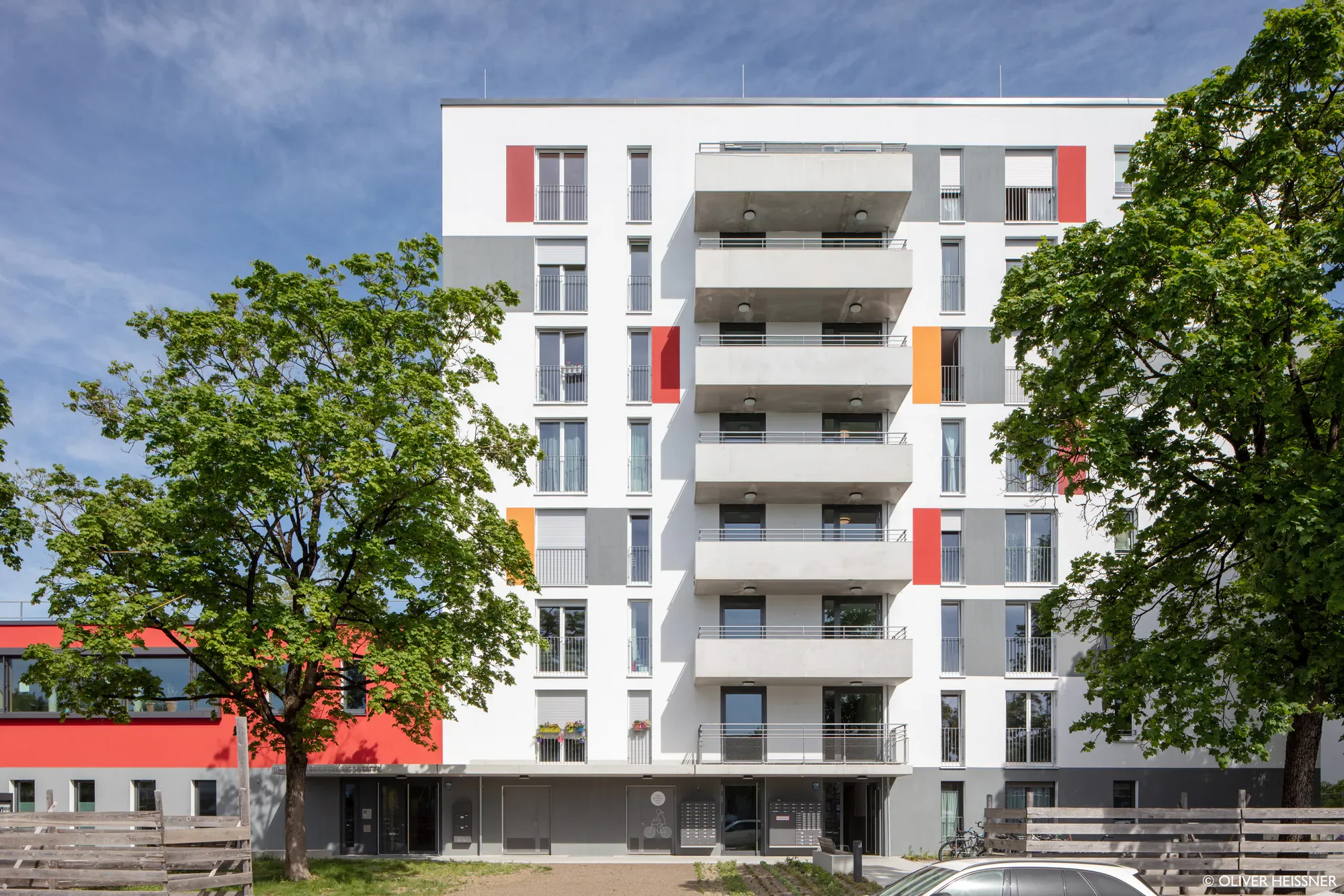 Mehrstöckiges modernes Apartmentgebäude mit weißen, grauen und orangefarbenen Akzenten, mehrere Balkone, umgeben von grünen Bäumen und einem blauen Himmel.