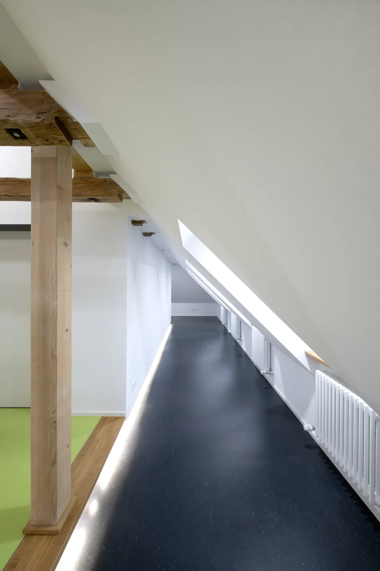 Innenraum mit schwarzem Boden, weißen Wänden und einem Dachfenster. Es gibt eine Heizkörperanordnung an der rechten Wand und eine Holzkonstruktion im Vordergrund.