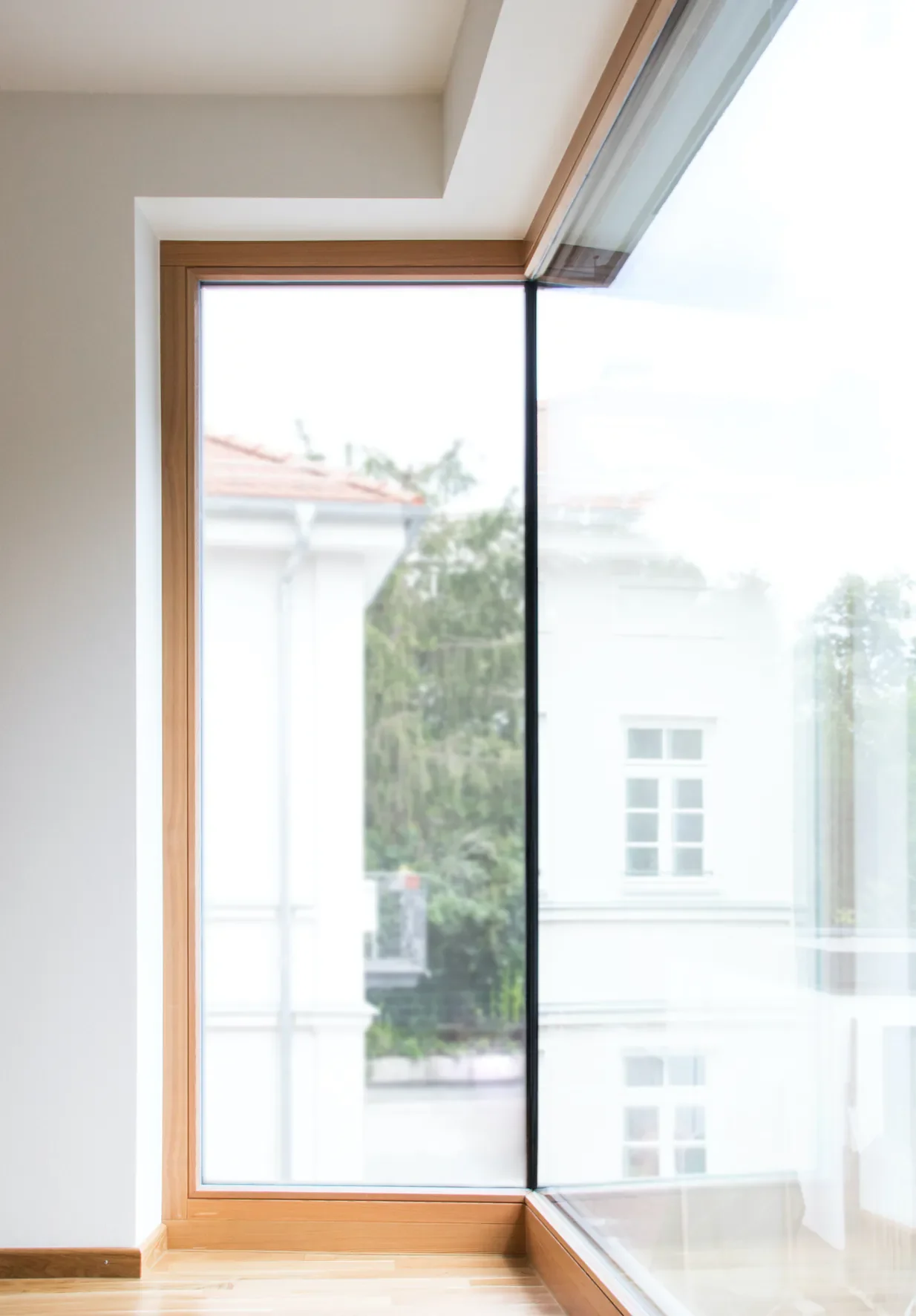 Großes Fenster mit Holzrahmen in einem modernen Raum, Blick nach draußen auf ein Gebäude und Bäume.