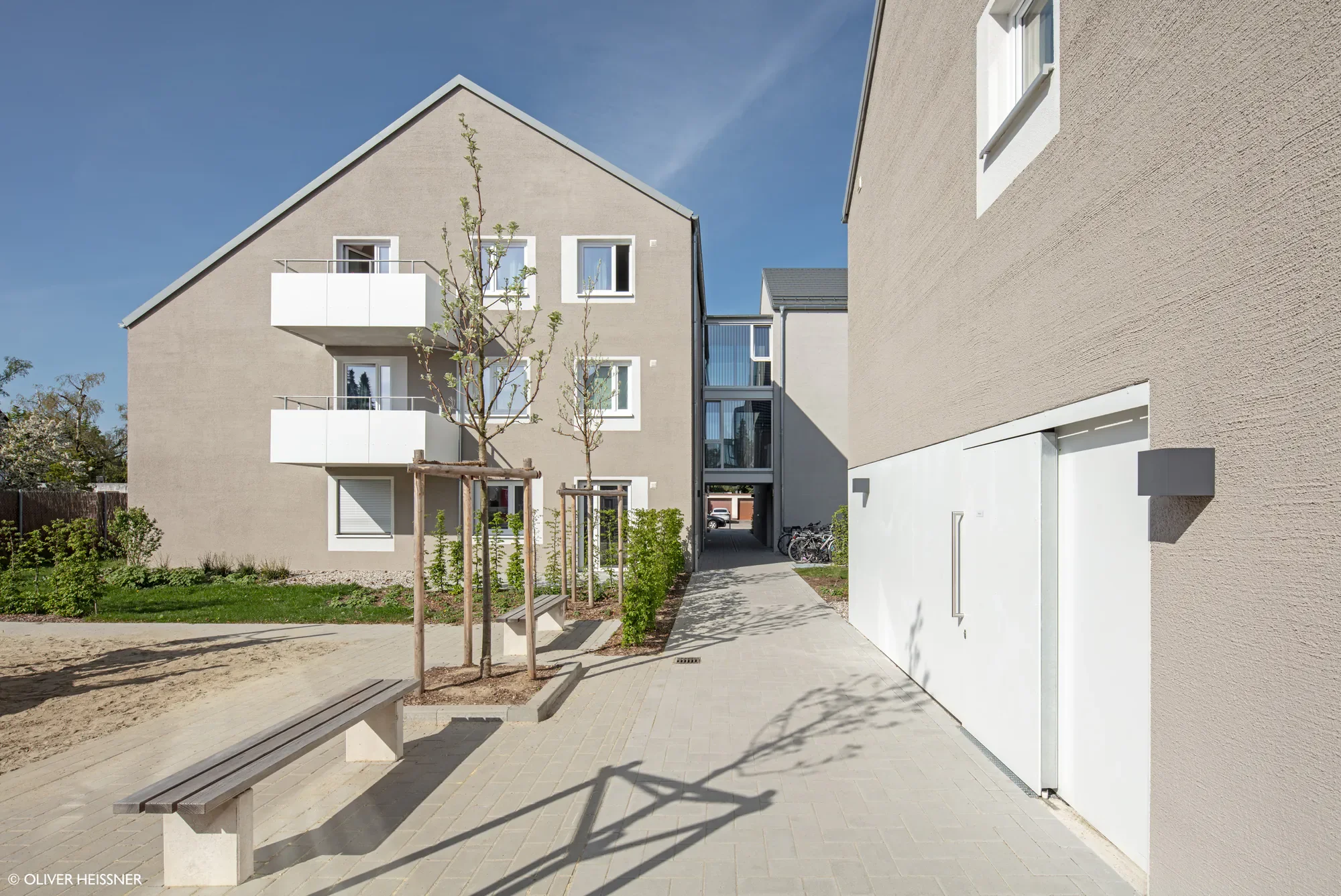 Moderne Mehrfamilienhaus mit grauer Fassade, Balkon und kleinen Bäumen im Vorgarten, Blick auf den hellen Gehweg und den Zugang zum Gebäude.