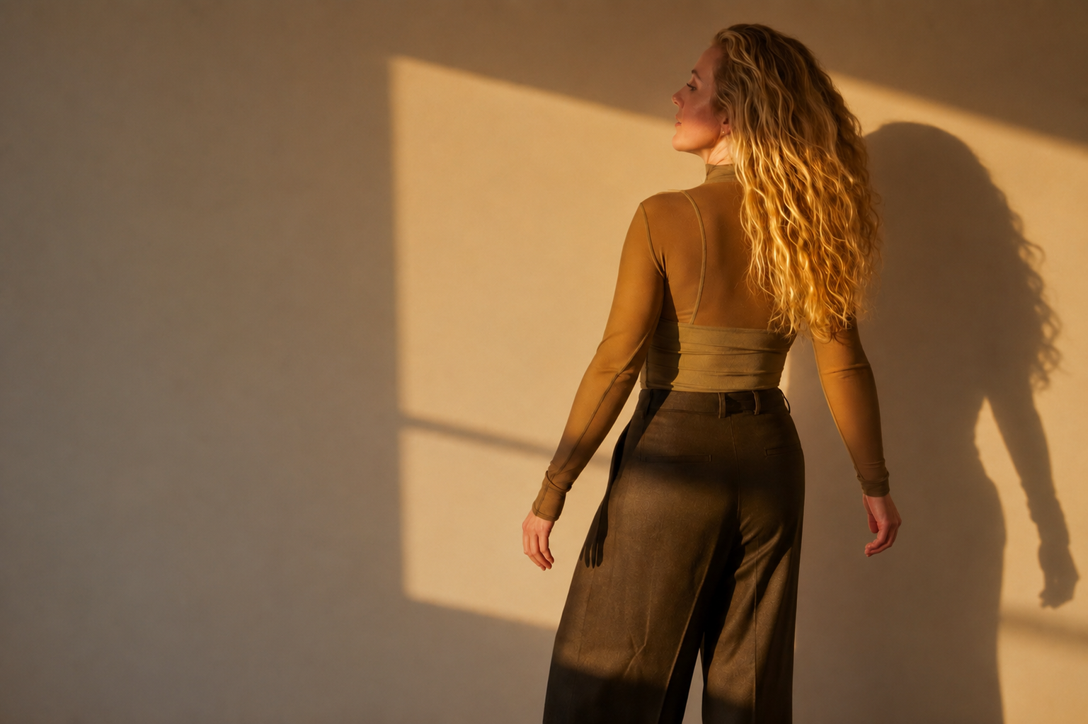 A woman with long, curly blonde hair stands against a beige wall illuminated by warm sunlight, casting a shadow of herself on the wall.