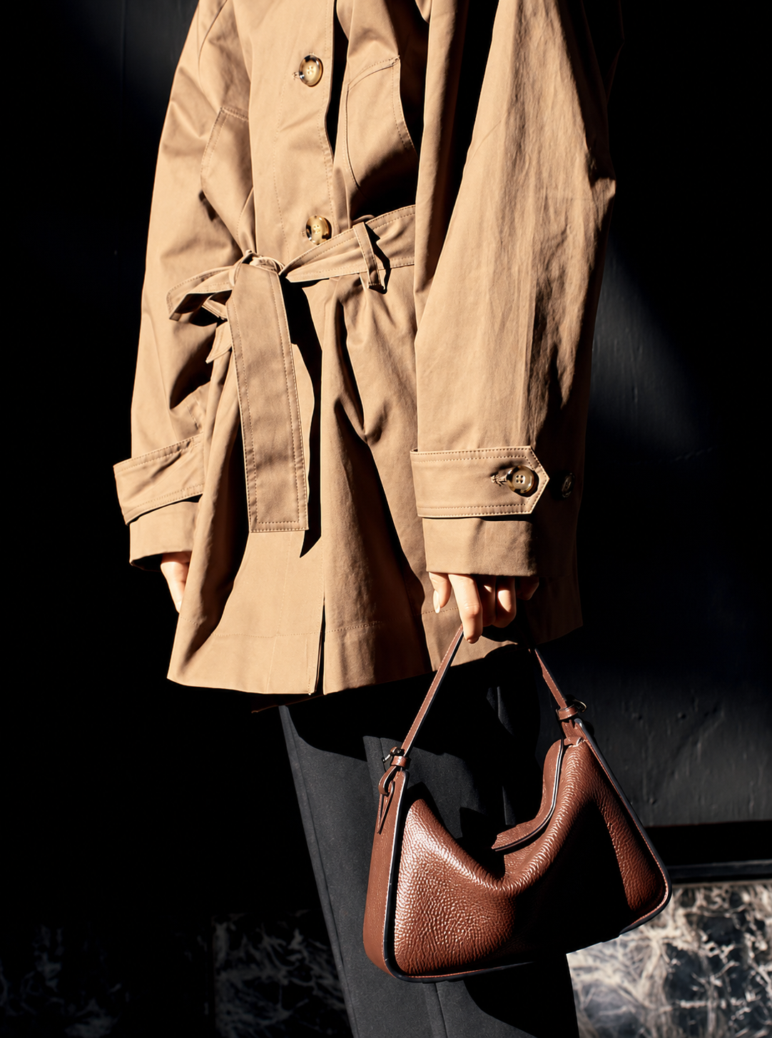 Close-up of a person wearing a beige trench coat, black pants, and holding a brown textured handbag, with the focus on the clothing and accessory against a dark background.