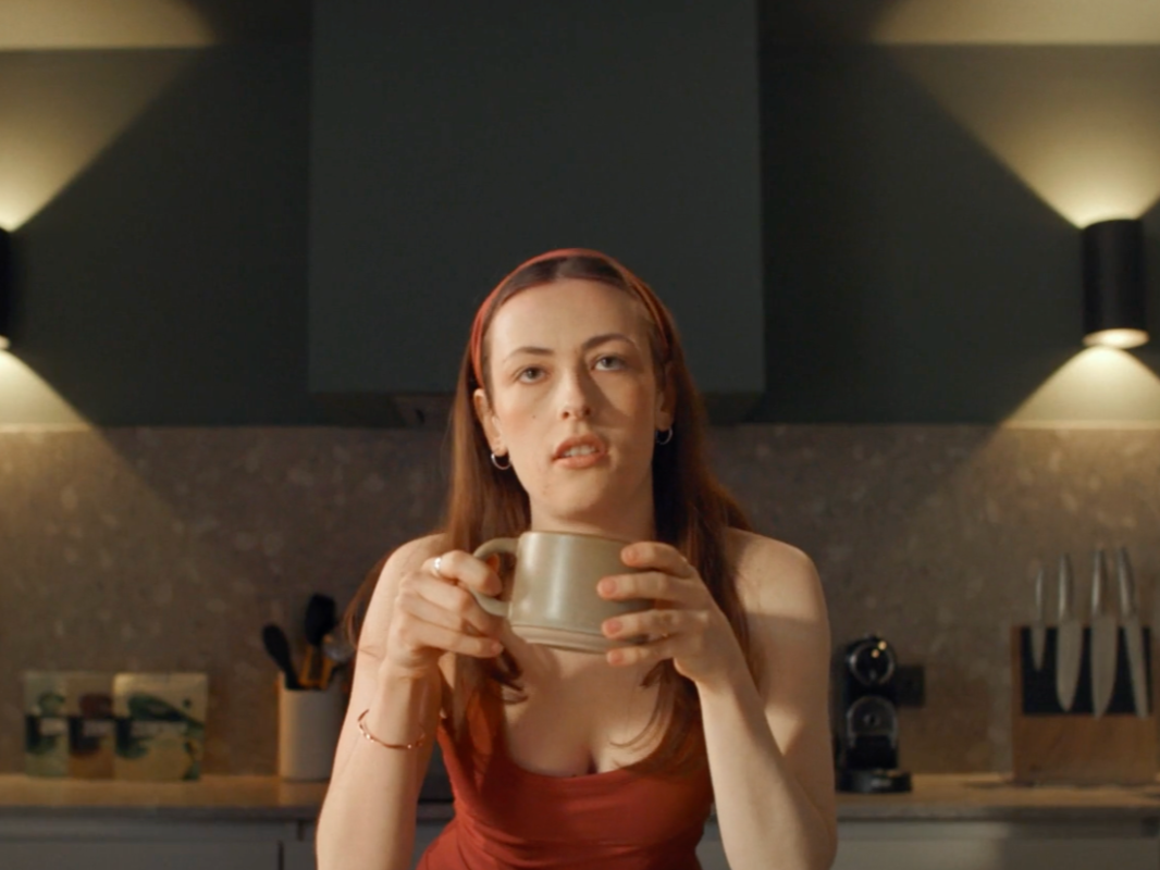 A woman with brown hair holding a coffee mug in a kitchen, looking at the camera with a neutral expression.