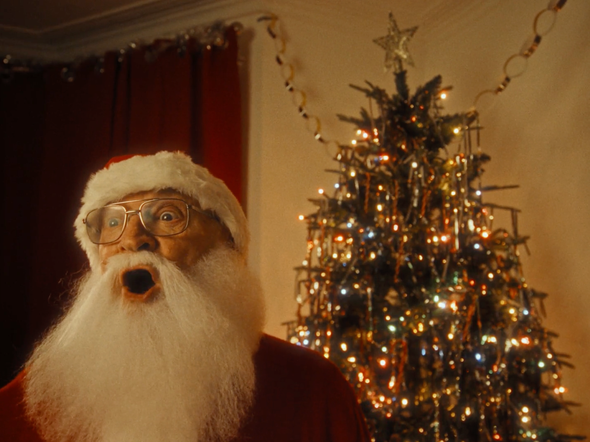 A person dressed as Santa Claus, with a Santa hat, glasses, and a white beard, standing in front of a decorated Christmas tree with colourful lights. The style of the set and costume is very retro.