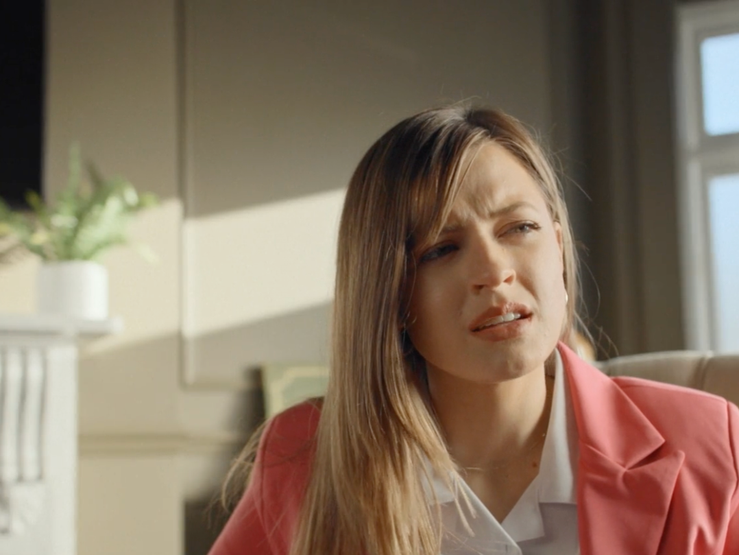 A woman with long, light brown hair wearing a pink blazer and white shirt sitting indoors near a window with sunlight shining on her, displaying a confused or questioning expression.