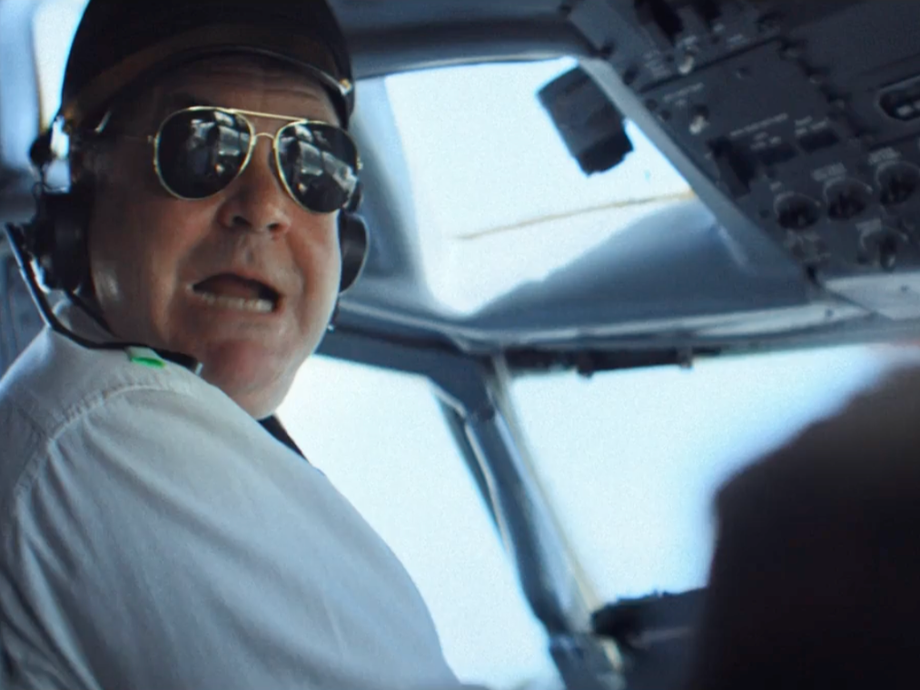 A pilot wearing sunglasses and headphones looking towards the camera inside an airplane cockpit.