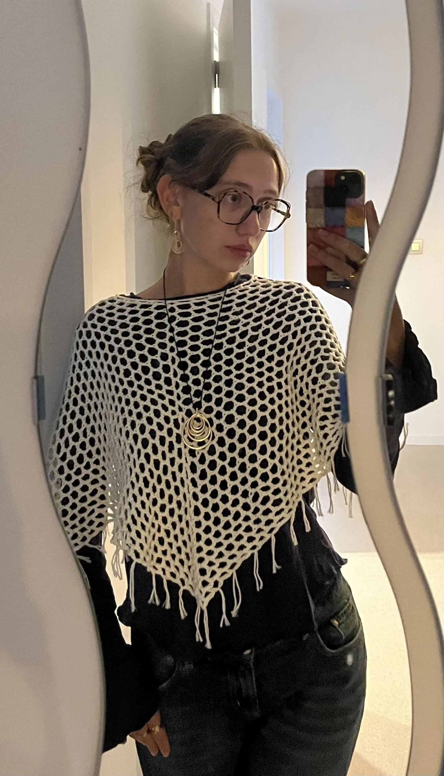 A young woman taking a mirror selfie in a room, wearing glasses, a black and white mesh top, large earrings, and a layered necklace, with her hair tied in a loose bun.