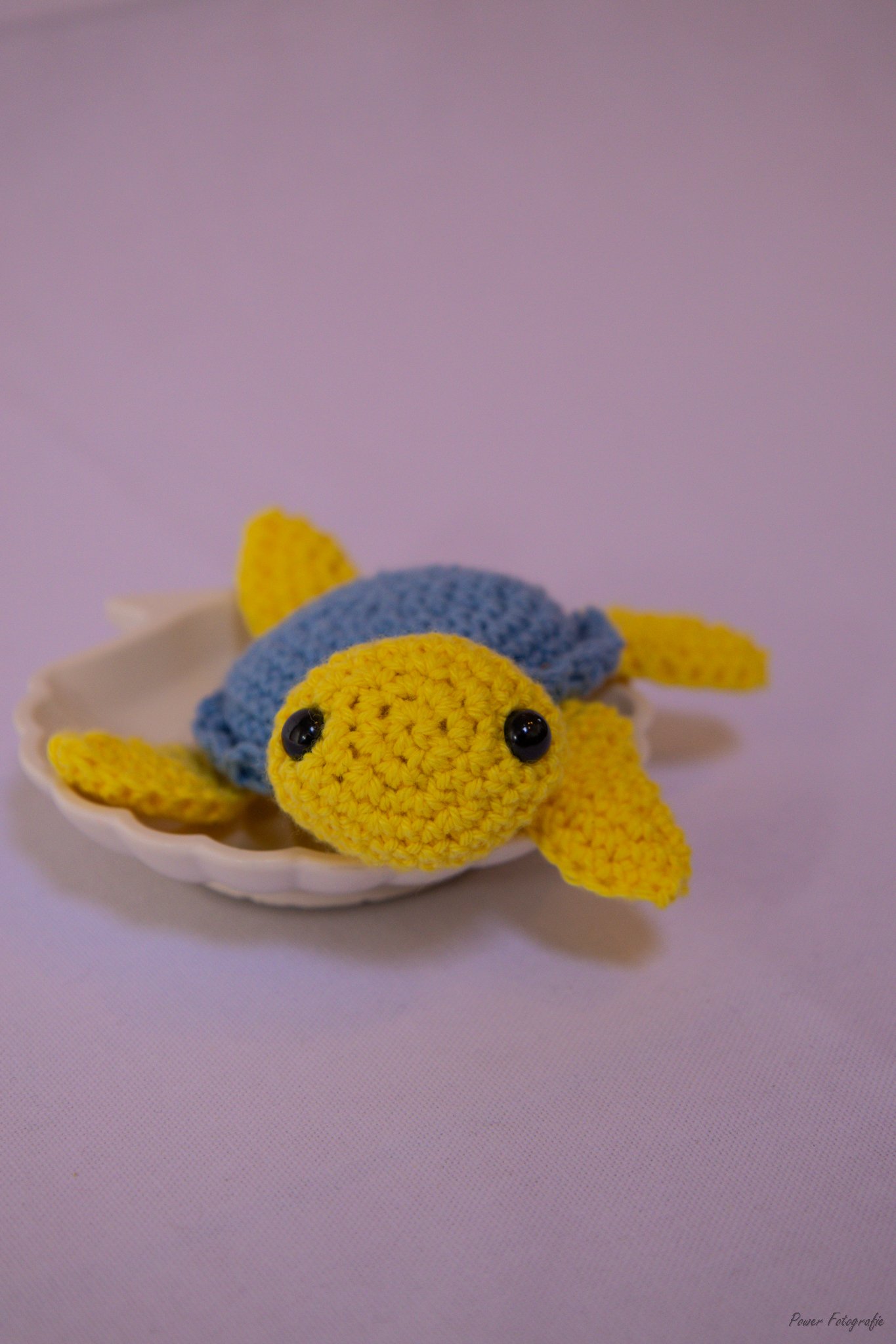 Handmade crocheted yellow and blue turtle plush toy on a white plate with a pink background.