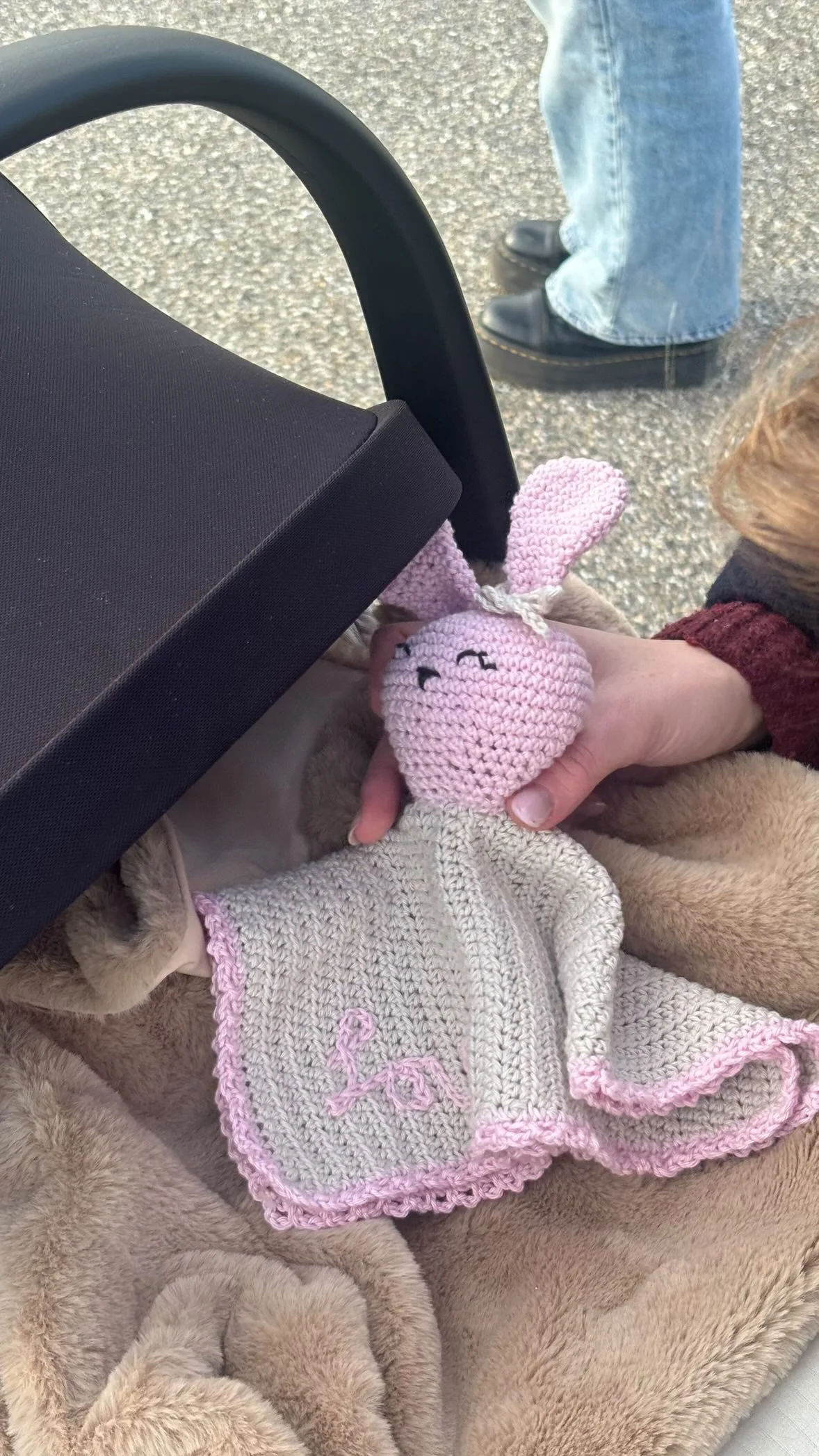 A person holding a pink crocheted bunny toy with embroidered face and large ears, dressed in a beige knit sweater with pink trim, resting on a fluffy beige blanket. Part of a wheelchair and other person's legs are visible in the background.