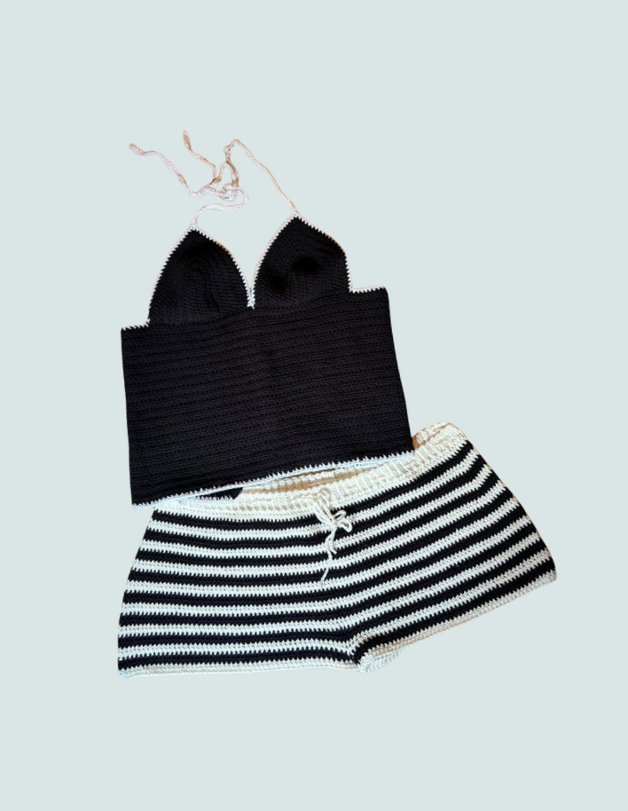 Black crochet bikini top with pink straps and striped black and white shorts laid flat on a light blue background.