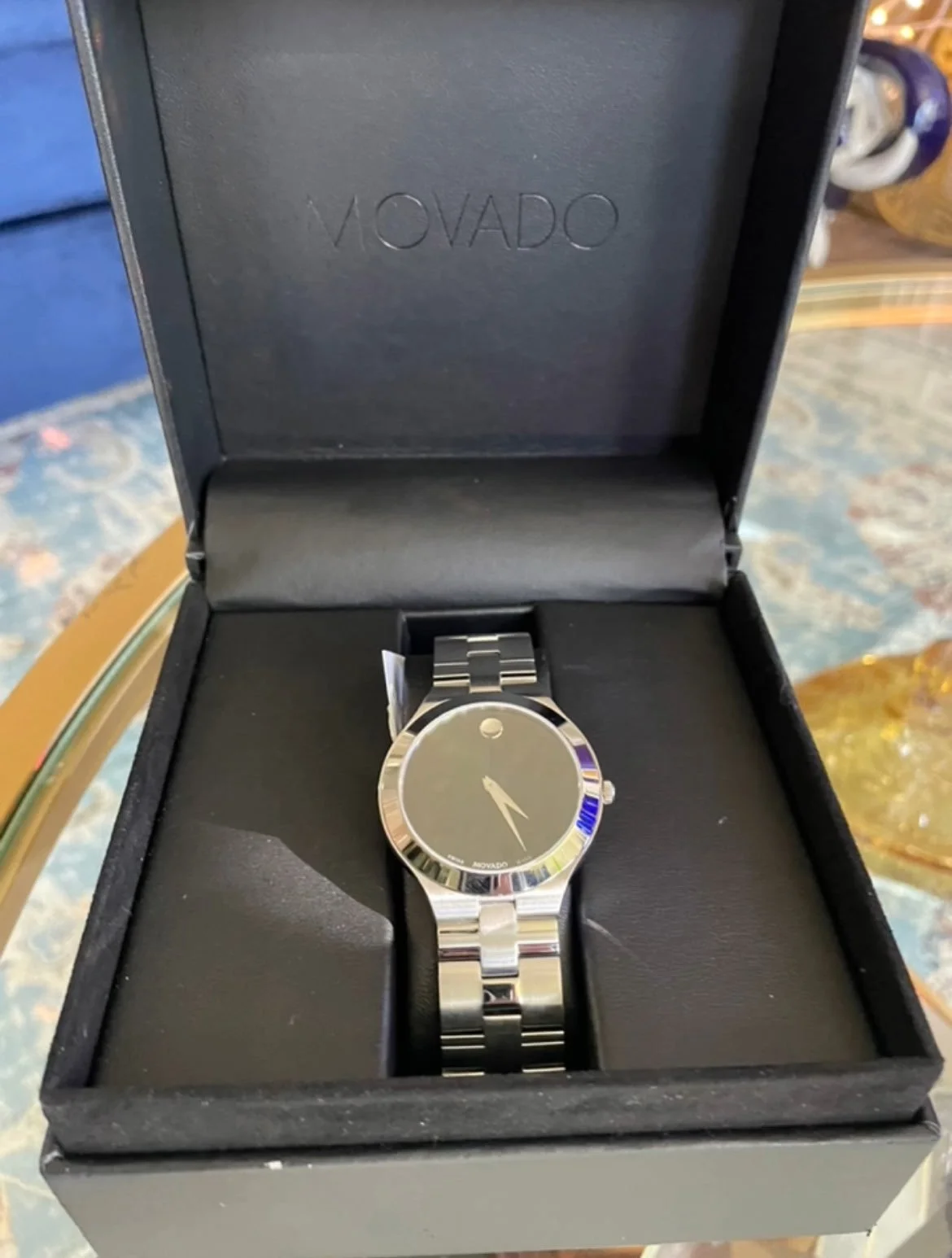 A silver Movado wristwatch with a black face and minimalist design, placed inside an open black box with the Movado branding on the inside lid.