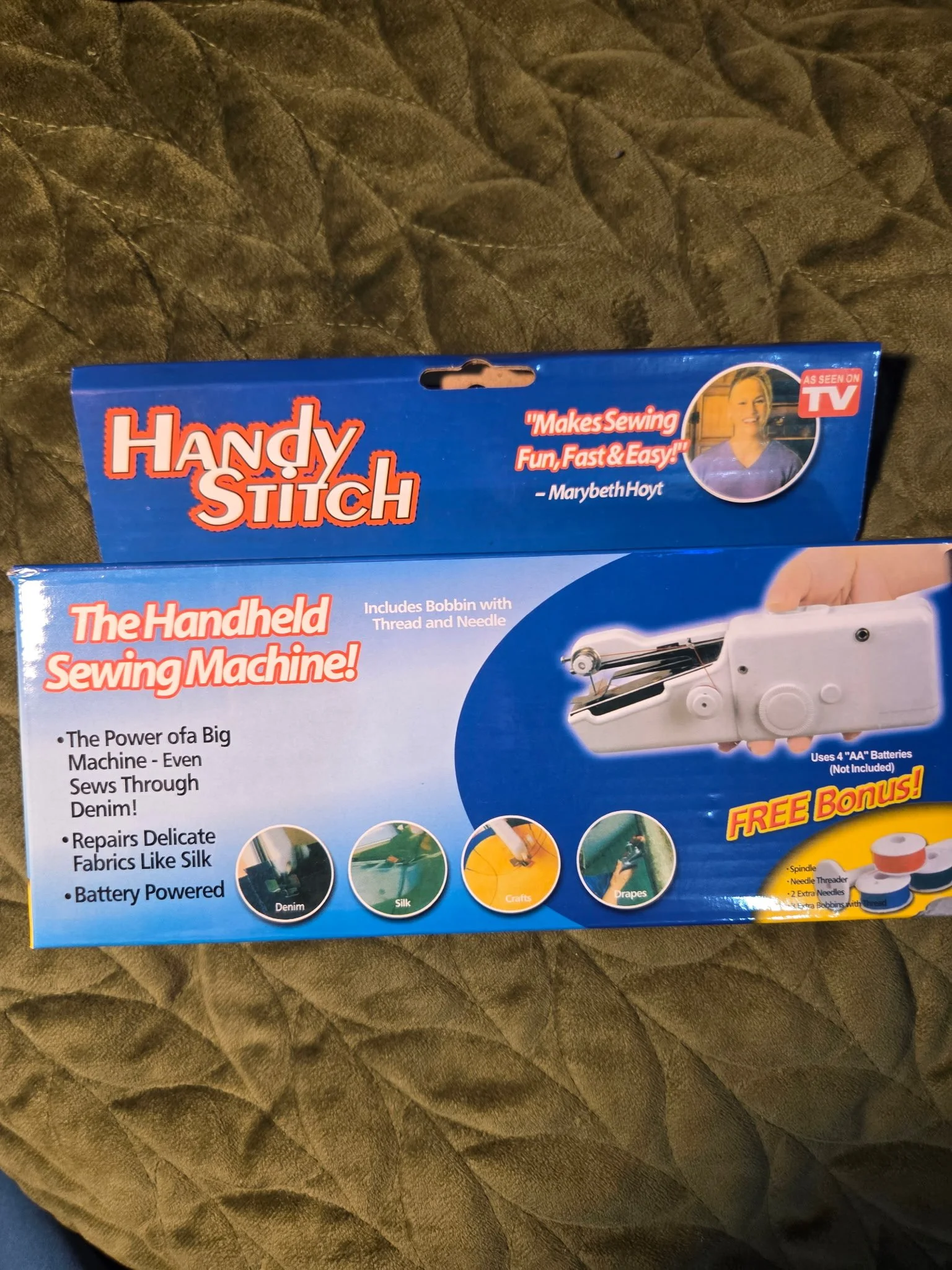 Blue packaging box for a handheld sewing machine named Handy Stitch. The box features an image of the sewing machine, a woman smiling, and text highlighting features like sewing through denim and silk, repairing delicate fabrics, and battery-powered operation. The box also shows small icons for denim, silk, crafts, and drapes.