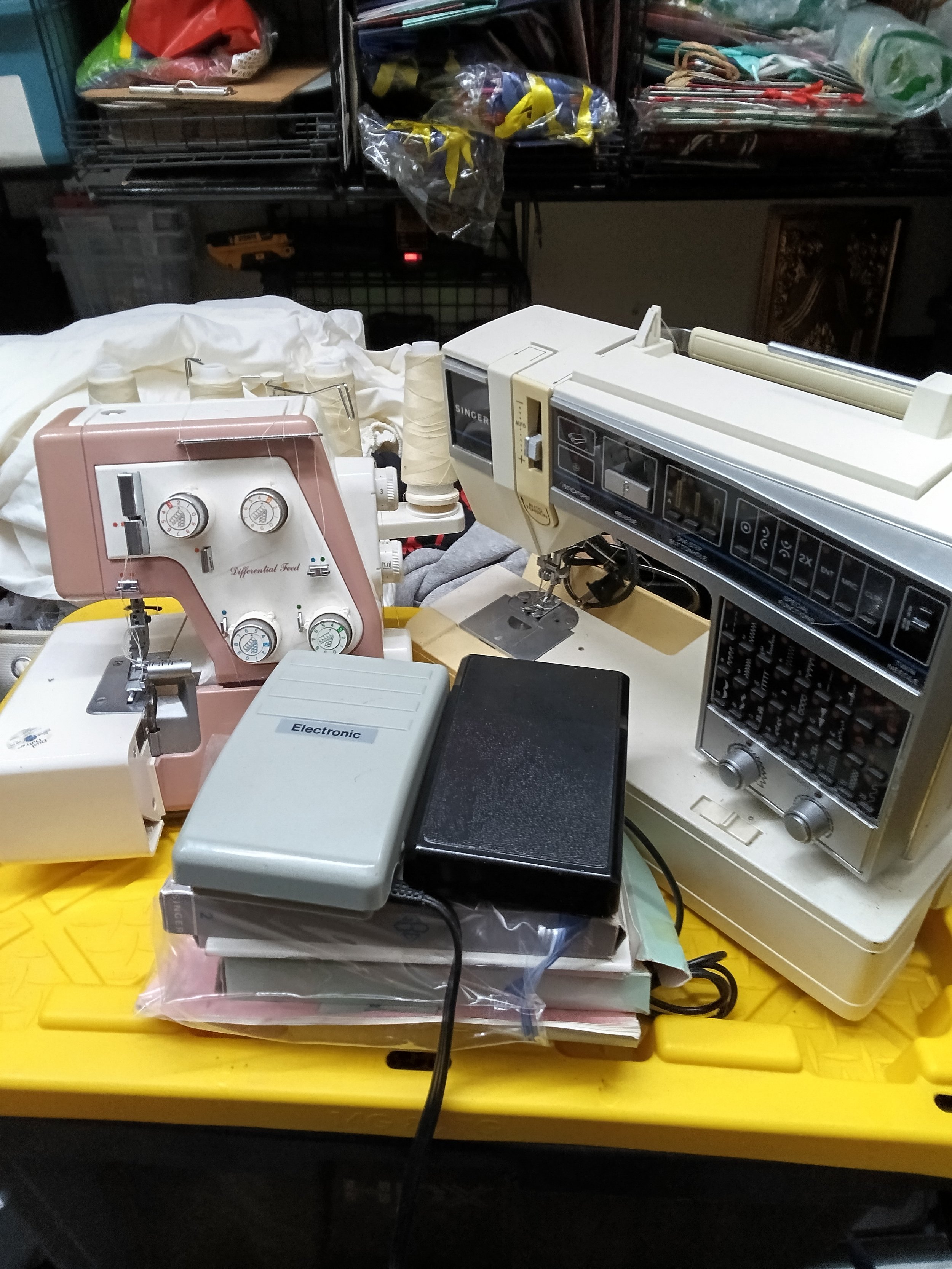 Sewing machine, cutting machine, and various sewing accessories on a yellow table in a cluttered workshop.