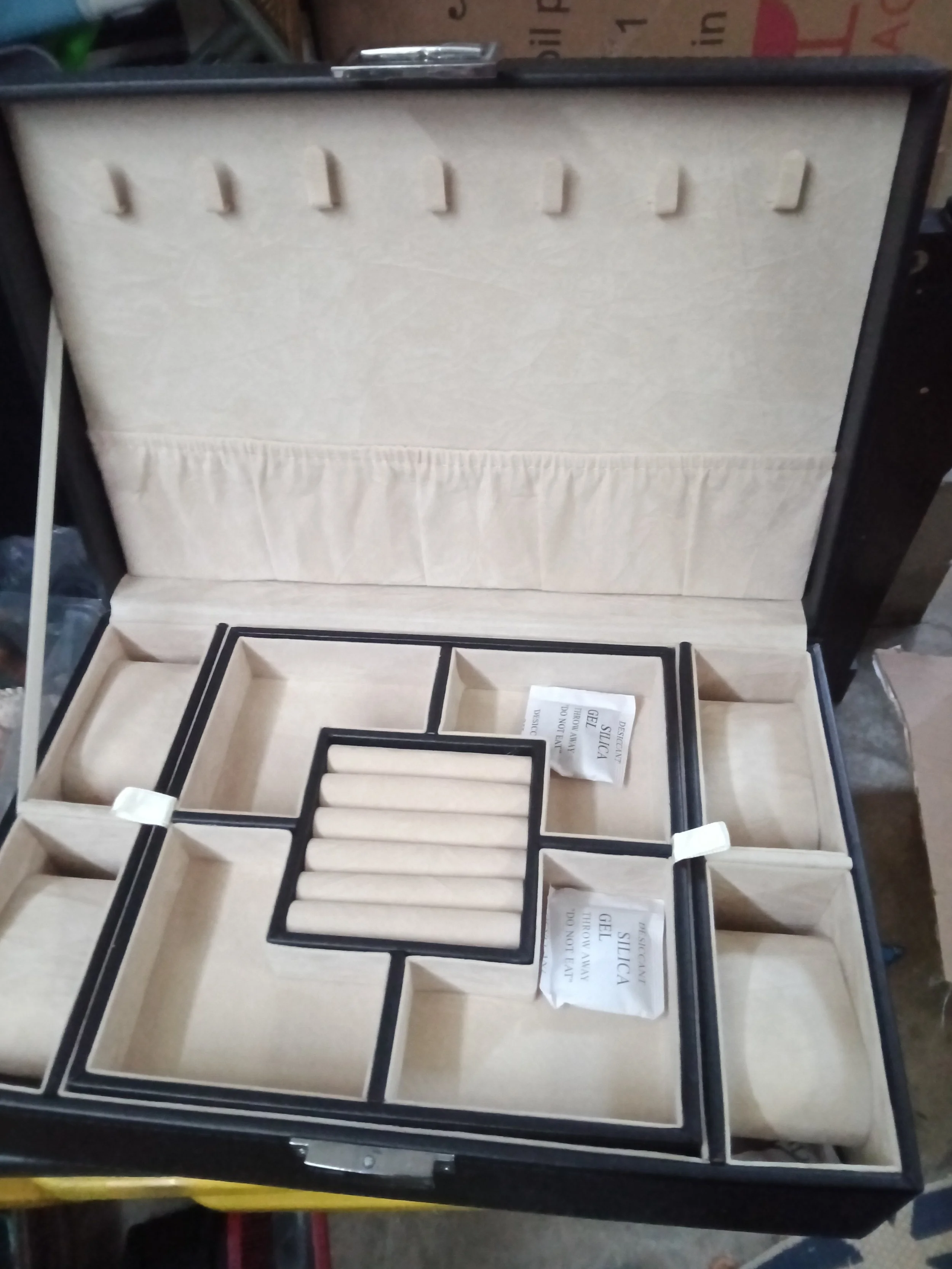 Open black jewelry or watch box with multiple compartments, lined with beige fabric, containing small foam insert holders. A paper label or instruction sheet is inside, but text is not fully visible.