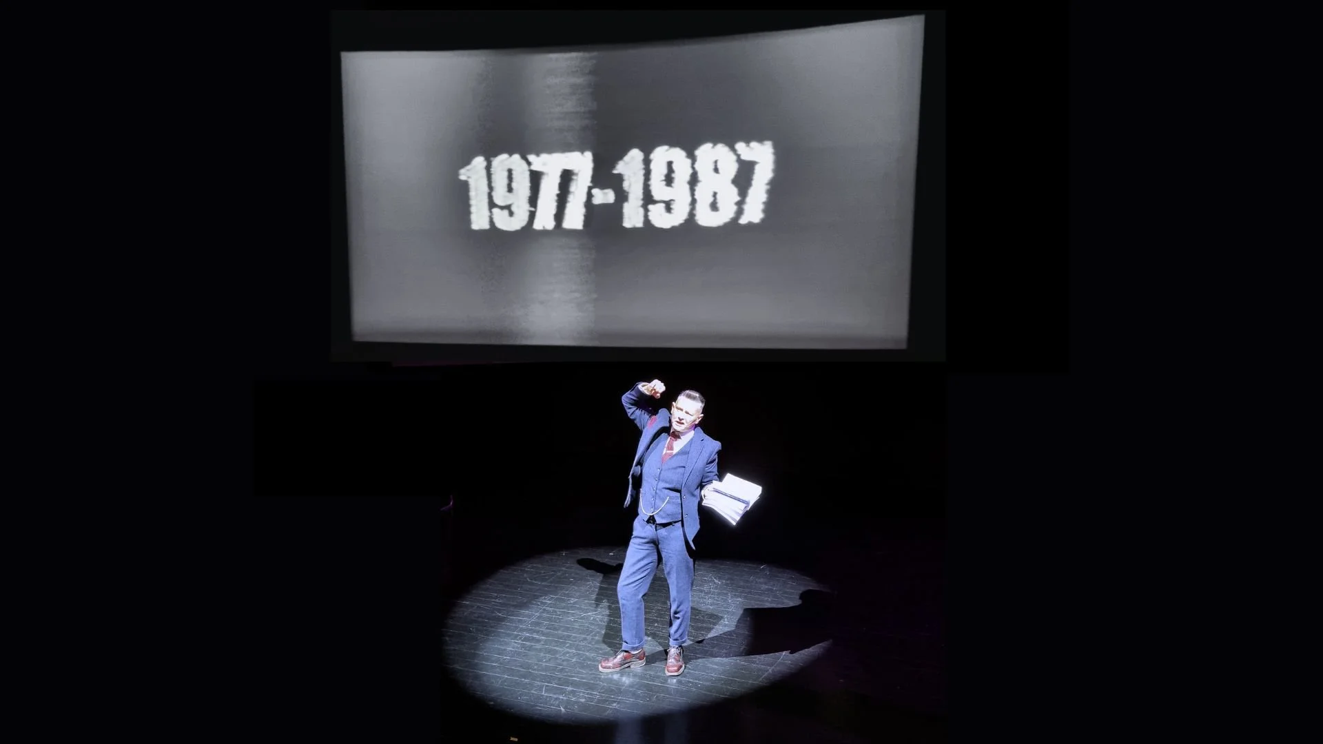 A person in a suit standing on a stage, gesturing with their fist raised, with a large screen behind them displaying the years '1977-1987'.