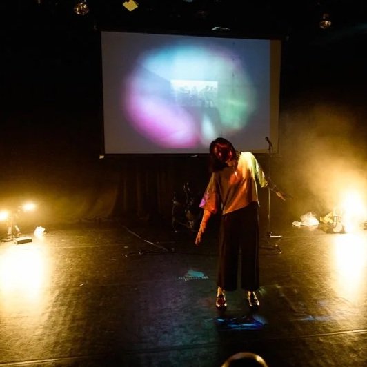 Person standing on stage with colorful lights and a blurred projection screen behind them.