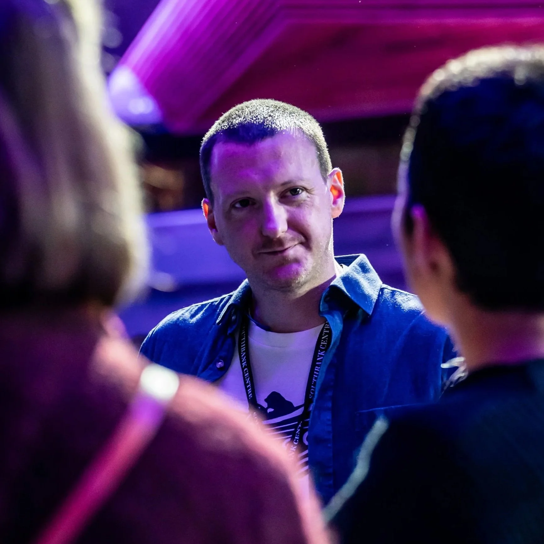 A group of people, with one man in focus, in a dimly lit environment with purple lighting. The man has short hair, is smiling slightly, and is wearing a blue shirt and a lanyard around his neck.