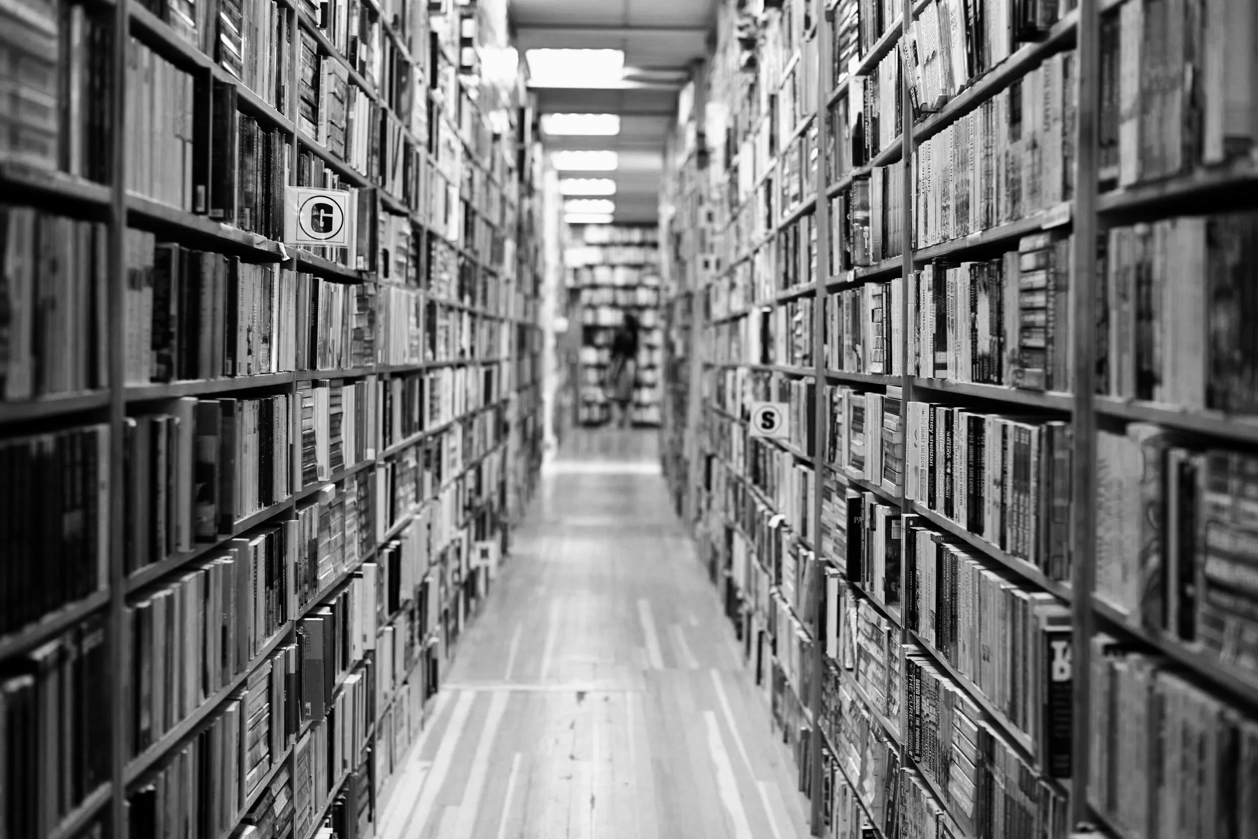 Black and white photo of tall bookshelves filled with books in a library aisle with signs indicating sections G and S.