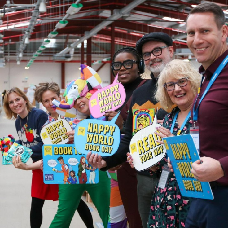 Group of people at a book event holding signs and books, celebrating World Book Day in a large indoor space.