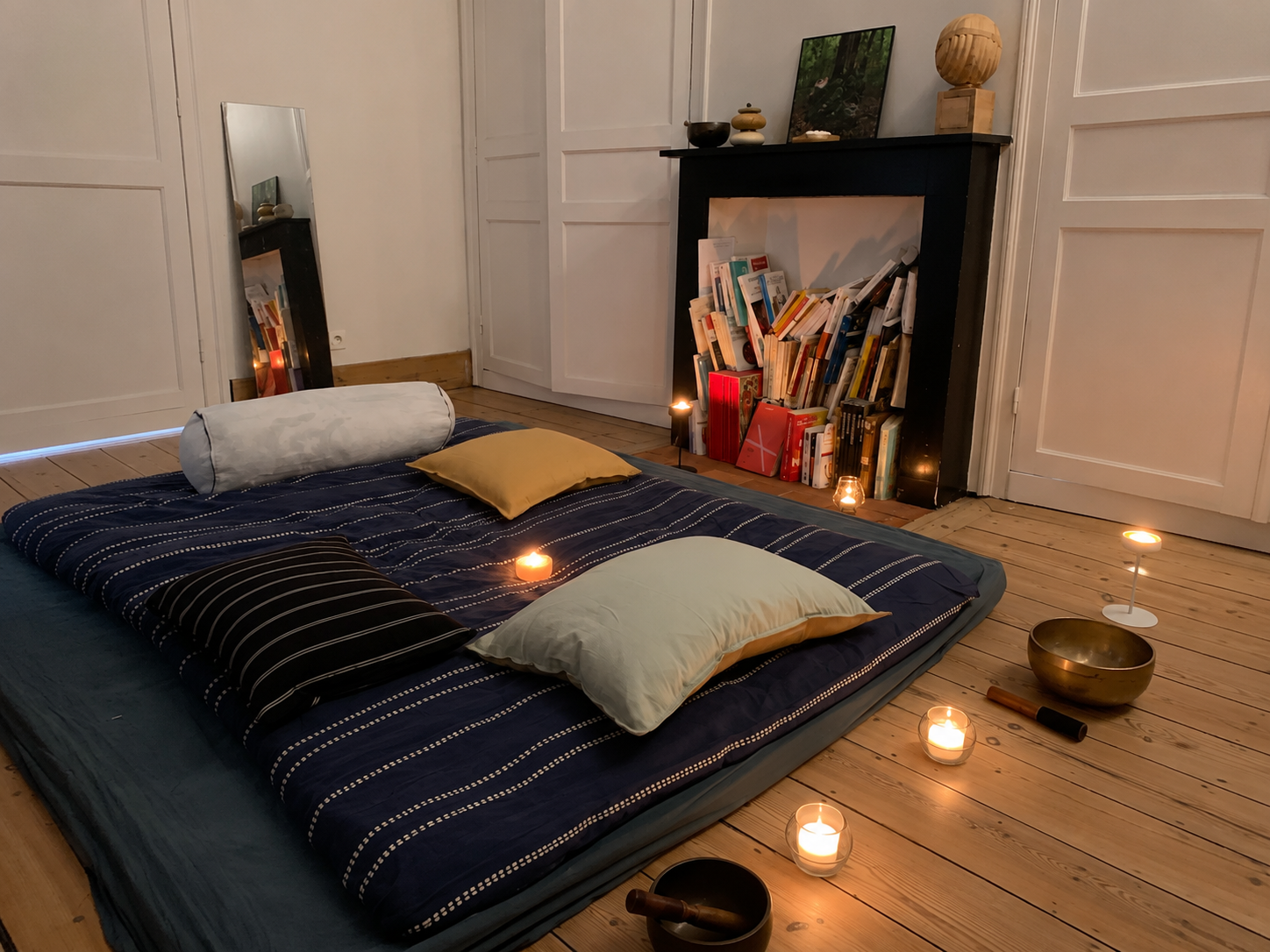 Une pièce chaleureuse avec un matelas de massage au sol, décorée avec des coussins confortables, des bougies relaxantes. Il y a une cheminée apaisante avec des livres. Endroit cosy et zen de massage à Lille.
