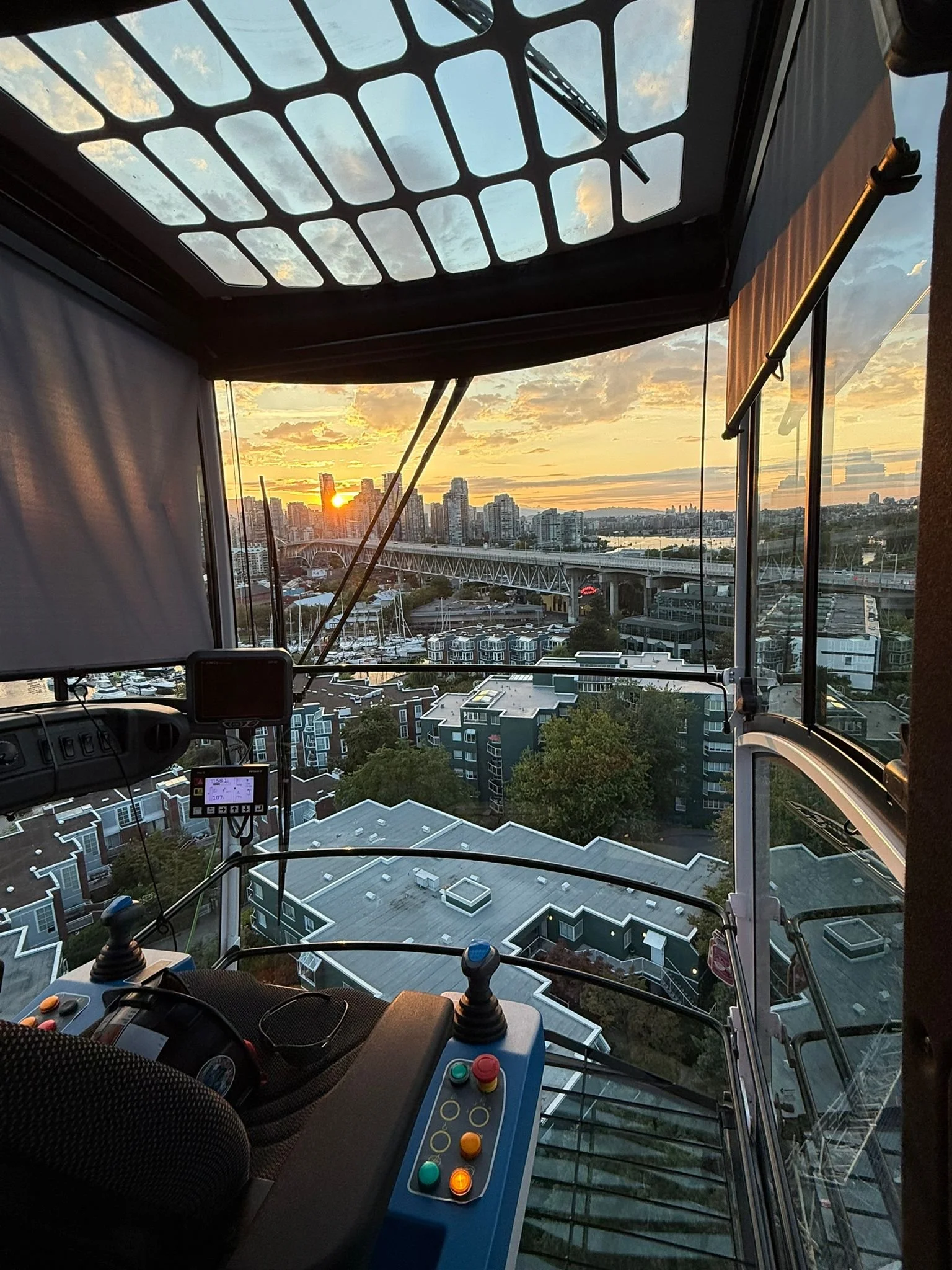 View from a glass elevator or observation pod showing a cityscape at sunset with buildings, a bridge, and a river.