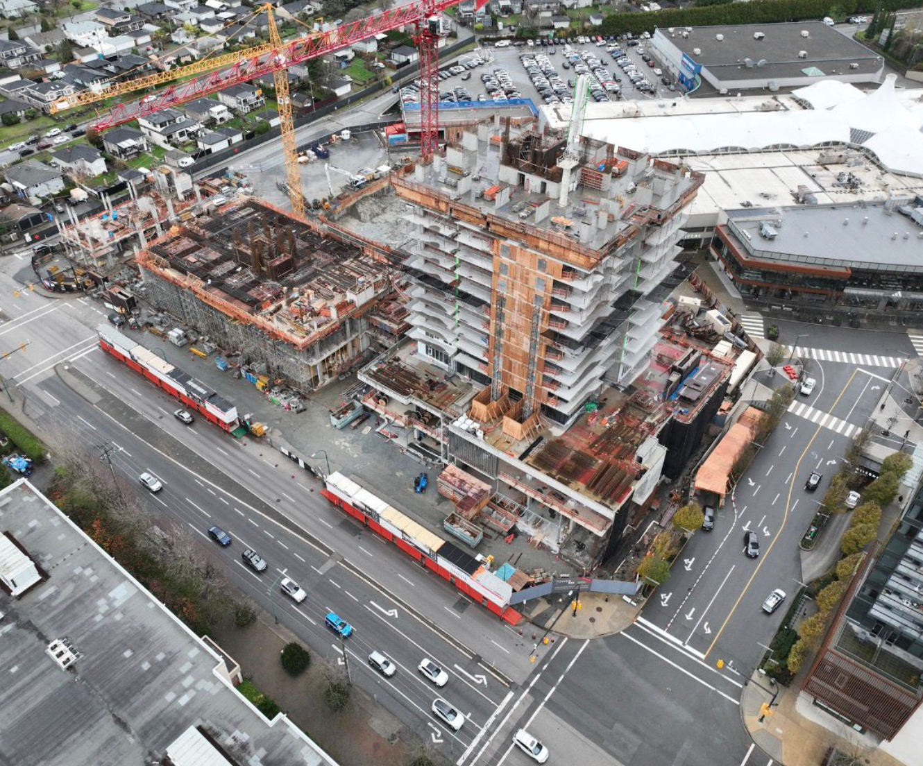 An aerial view of a construction site with a partially built high-rise building, cranes, and construction equipment, surrounded by roads and nearby buildings.