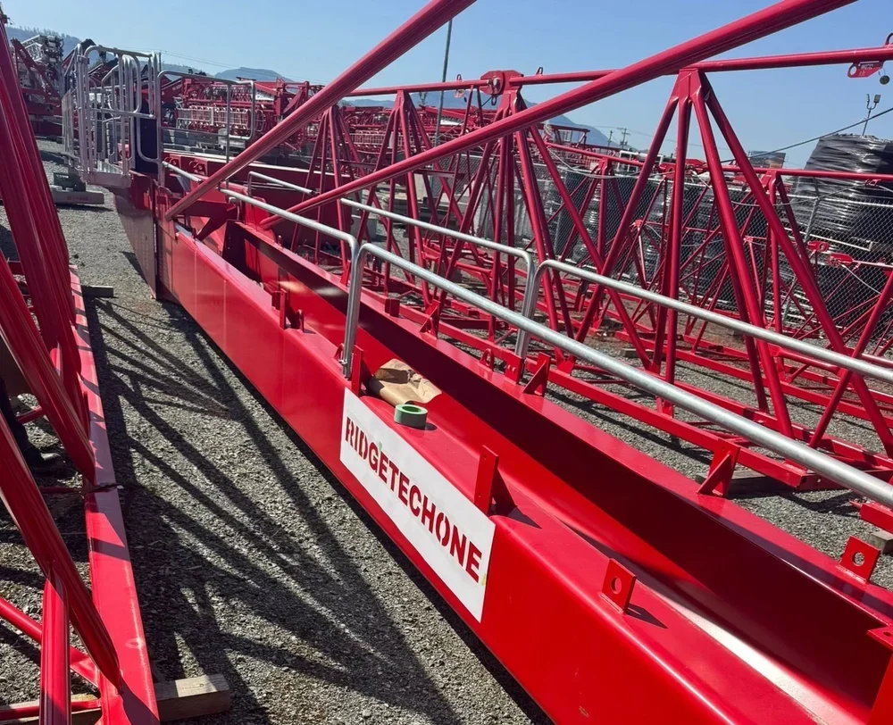 A red metal amusement park ride structure labeled "HUGGETCHONE," with various interconnected metal beams and safety railings, situated outdoors on a gravel lot with other similar structures in the background.