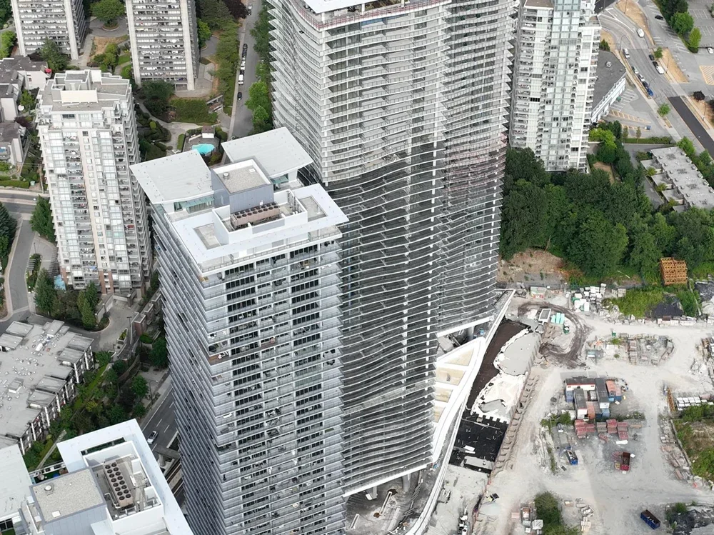 Aerial view of a modern high-rise building with a wavy glass facade amidst other tall buildings and a wooded area in a cityscape.