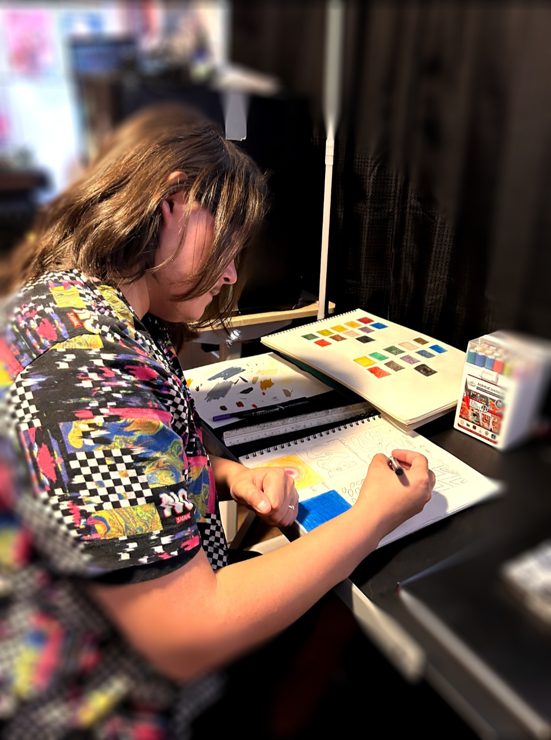 A man with shoulder-length brown hair, wearing a colorful patterned shirt, sitting at a desk and drawing or coloring in a sketchbook.
