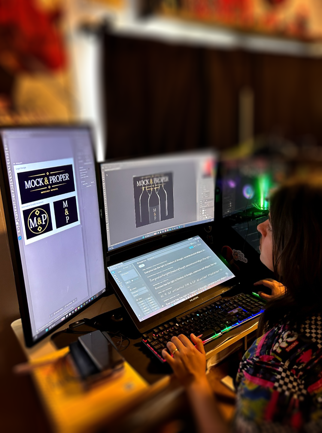 A person working at a computer designing a logo for 'Mock & Proper' with three different logo concepts displayed on three monitors.