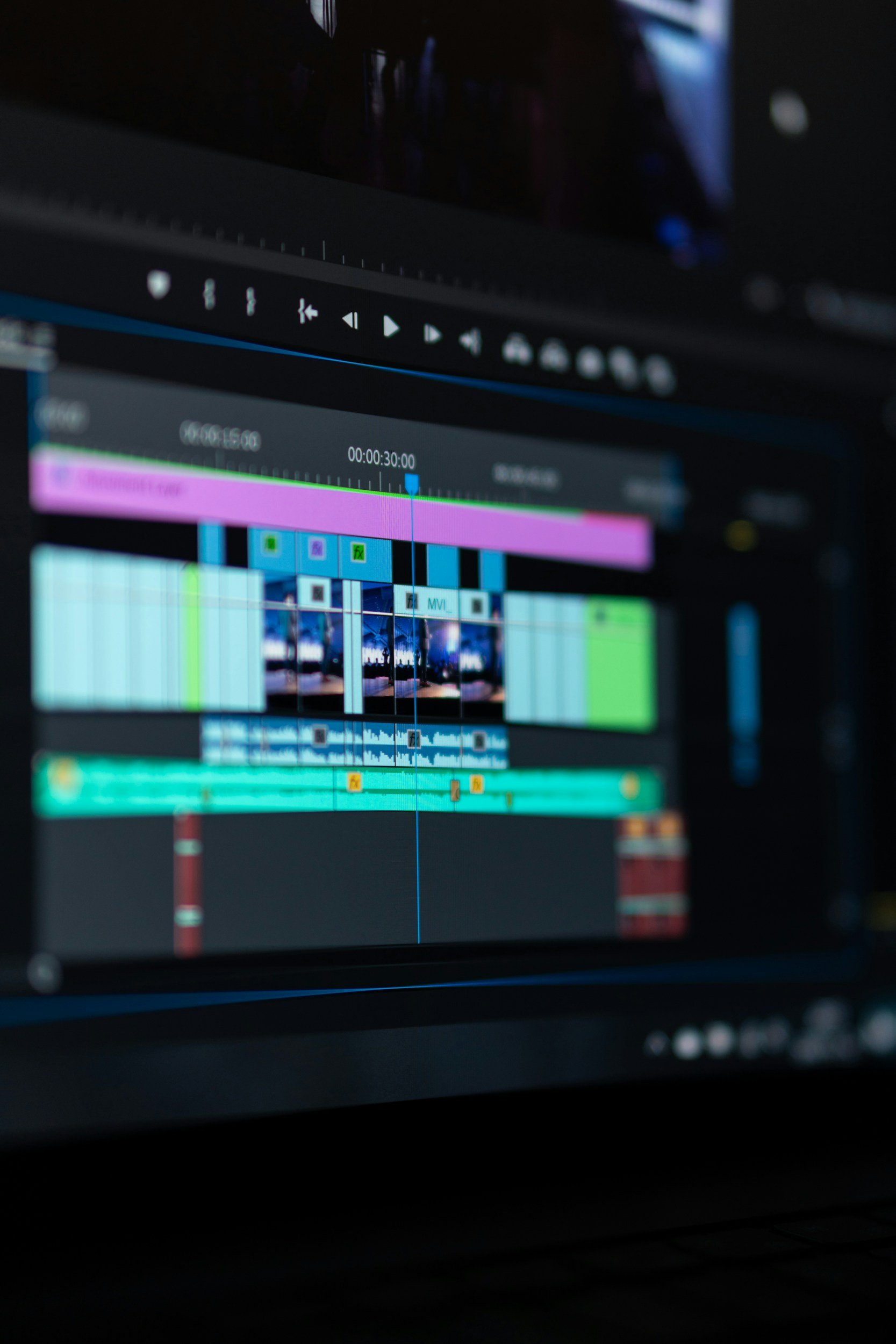 A computer screen displaying a video editing timeline with various video and audio tracks, playback controls at the top, and a dark background.