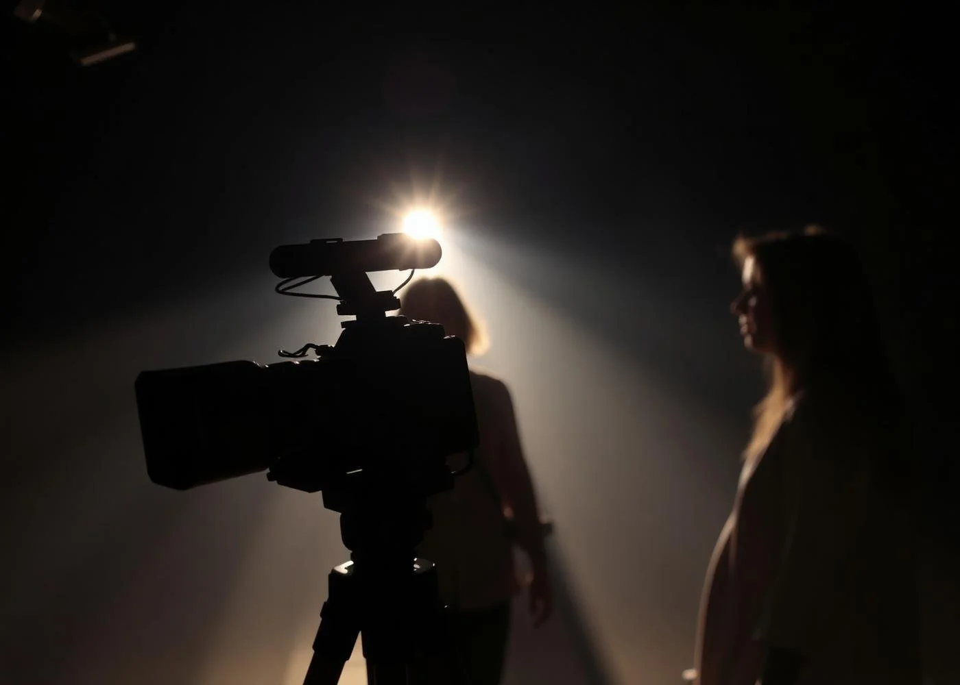 Silhouette of two women in a dark room with a spotlight, one woman is in front of a camera on a tripod, the other woman is slightly behind and to the right, both are facing different directions.