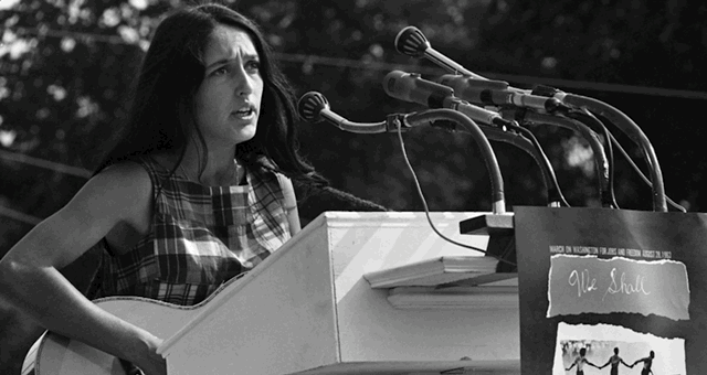 A woman speaking at a podium with multiple microphones, wearing a plaid dress in an outdoor setting.