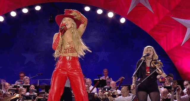 Two female performers on stage with an orchestra, one singing into a microphone and the other playing a violin during a patriotic themed event.