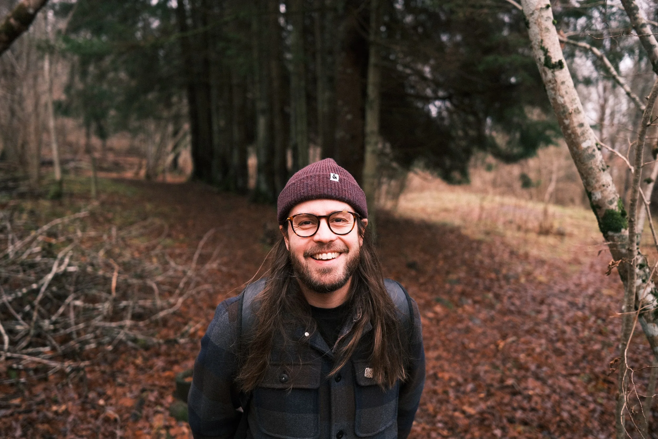 A man with long hair, glasses, and a beard wearing a maroon beanie and black jacket, smiling outdoors in a wooded area with fallen leaves and trees.