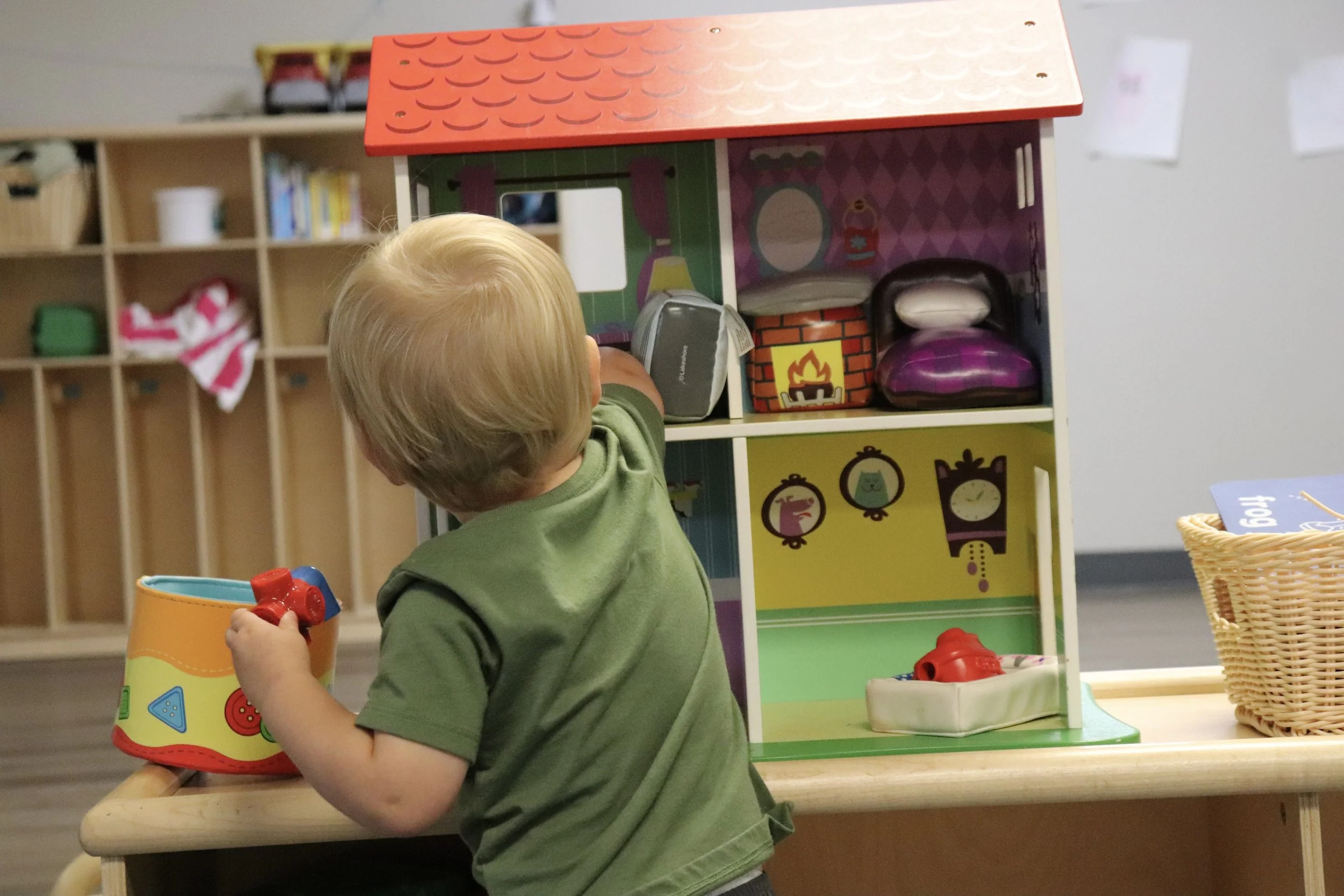 A young child with blond hair, wearing a green shirt, playing with a colorful toy bucket in a classroom or play area.