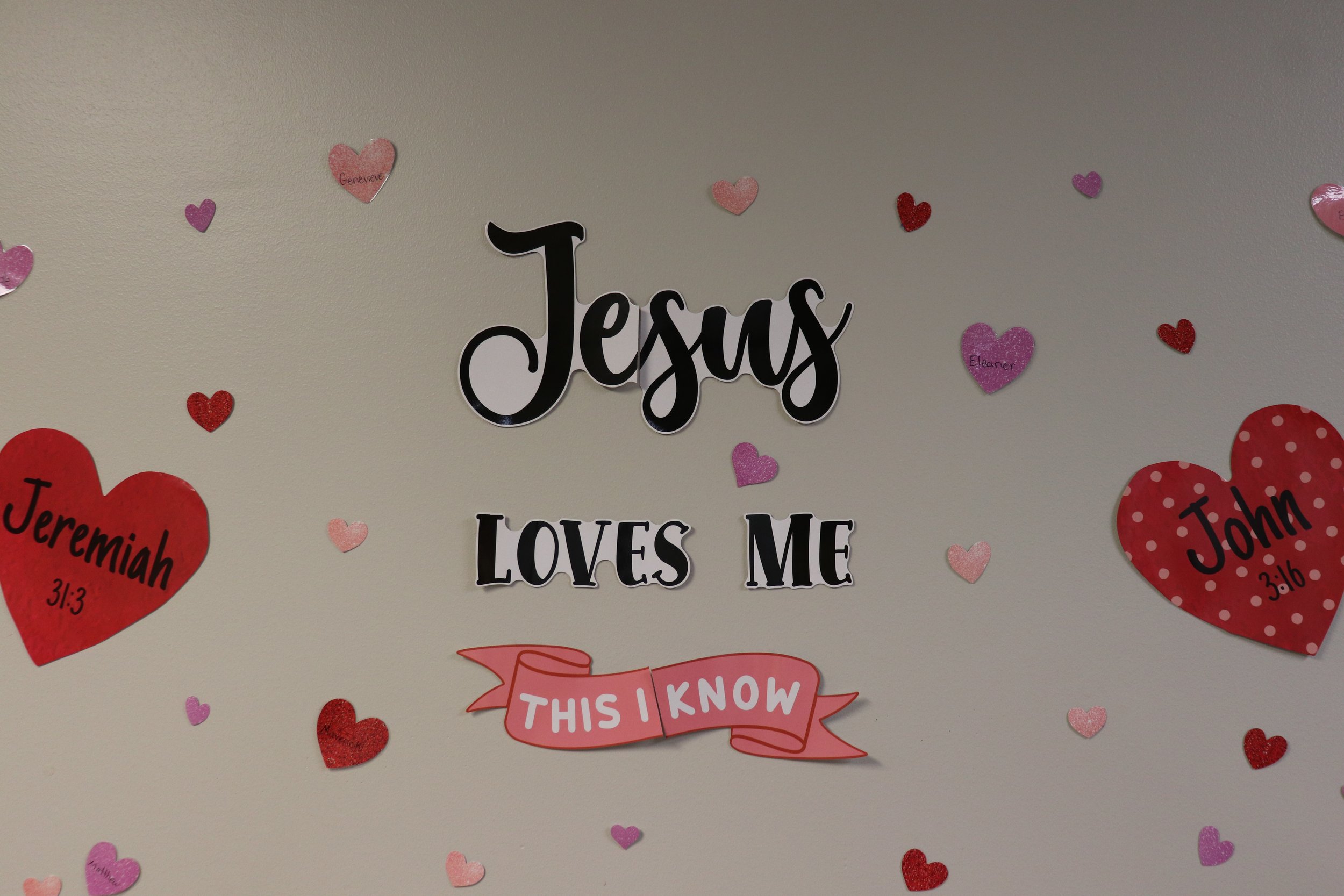 Decorative wall with the word 'Jesus' and the phrase 'loves me' surrounded by pink and red glitter hearts, some labeled with names and Bible verses.