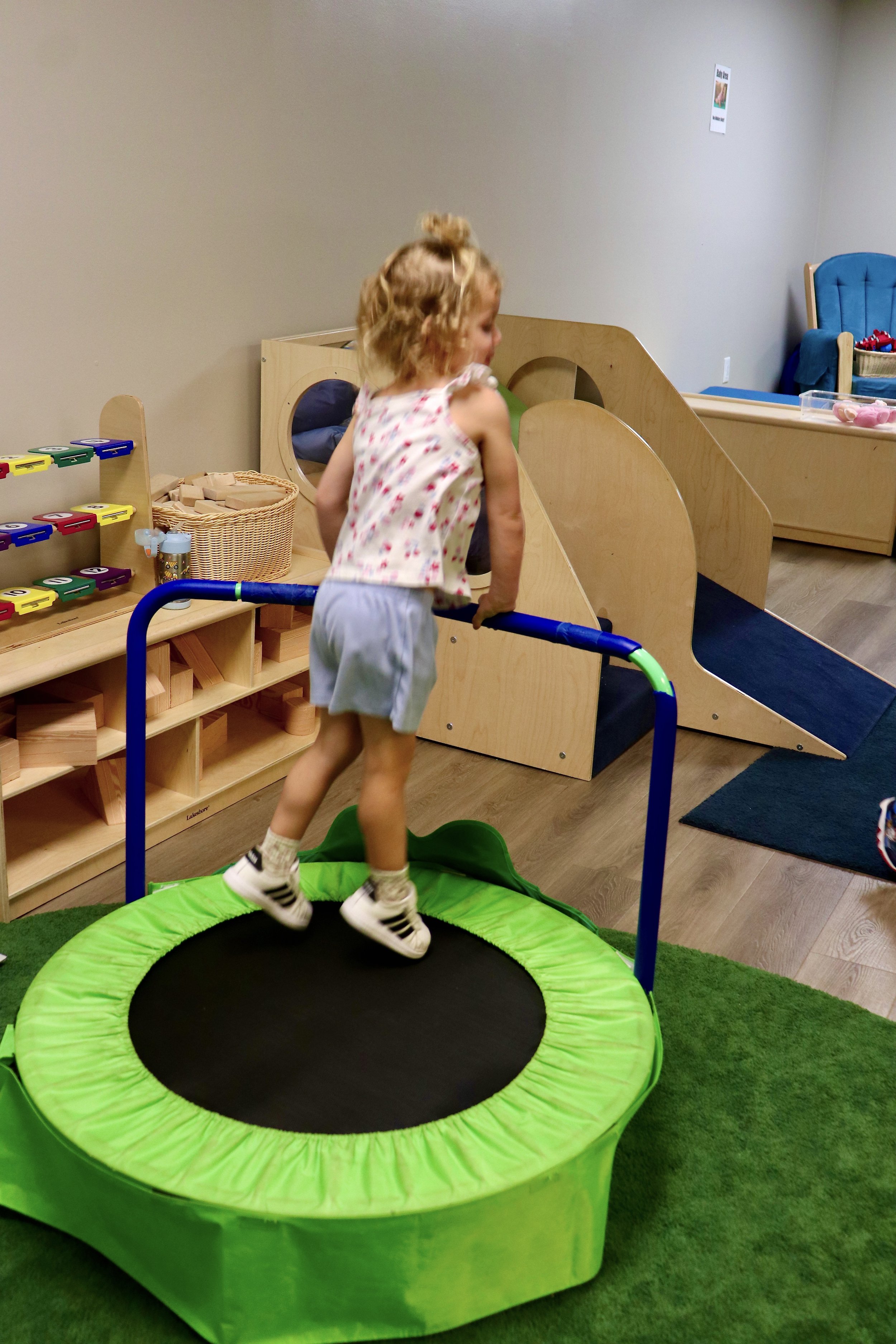 A young girl with curly hair jumping on a small green trampoline with black surface in a playroom.