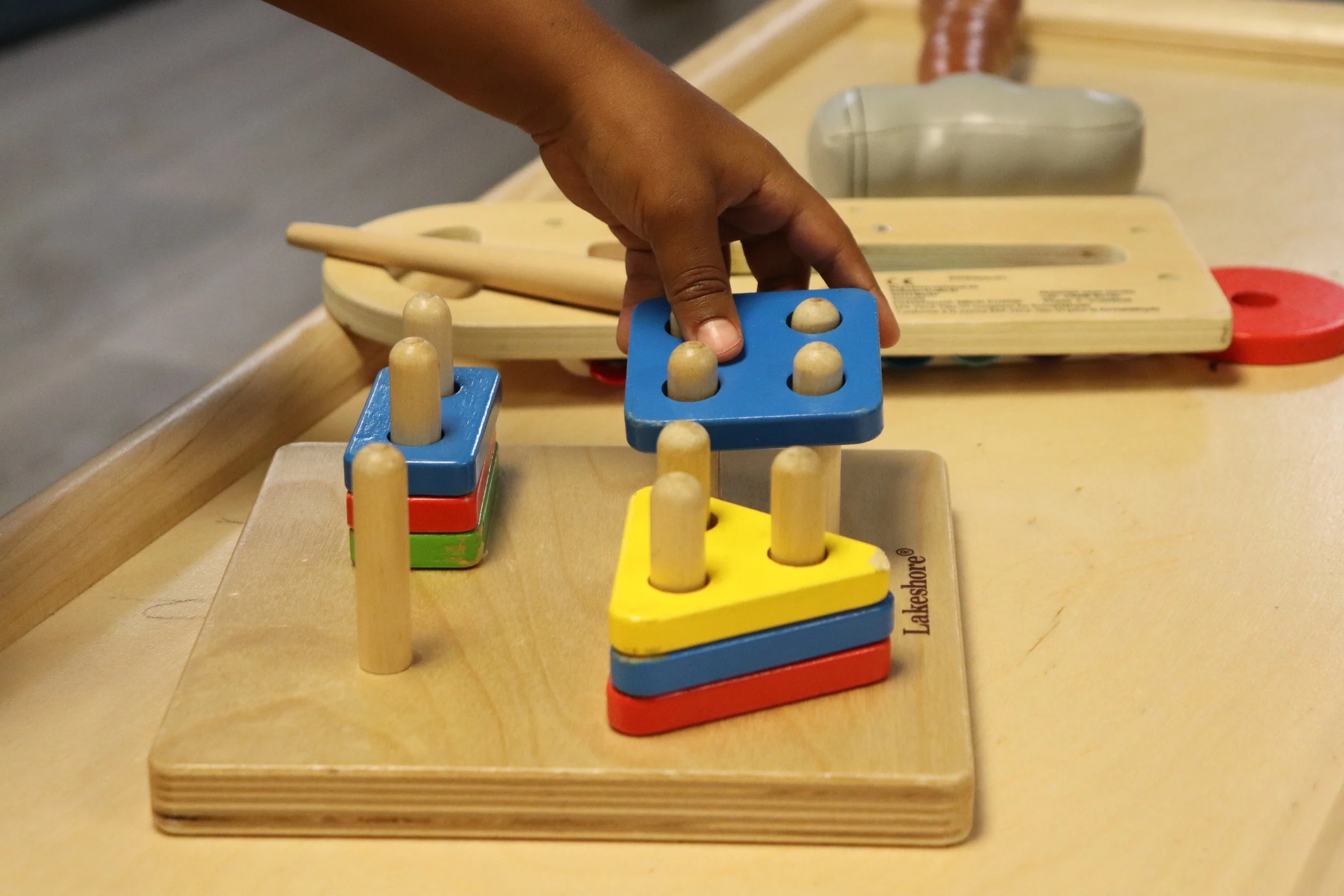 Child's hand playing with colorful wooden stacking toy and a wooden shape-sorting toy on a table.