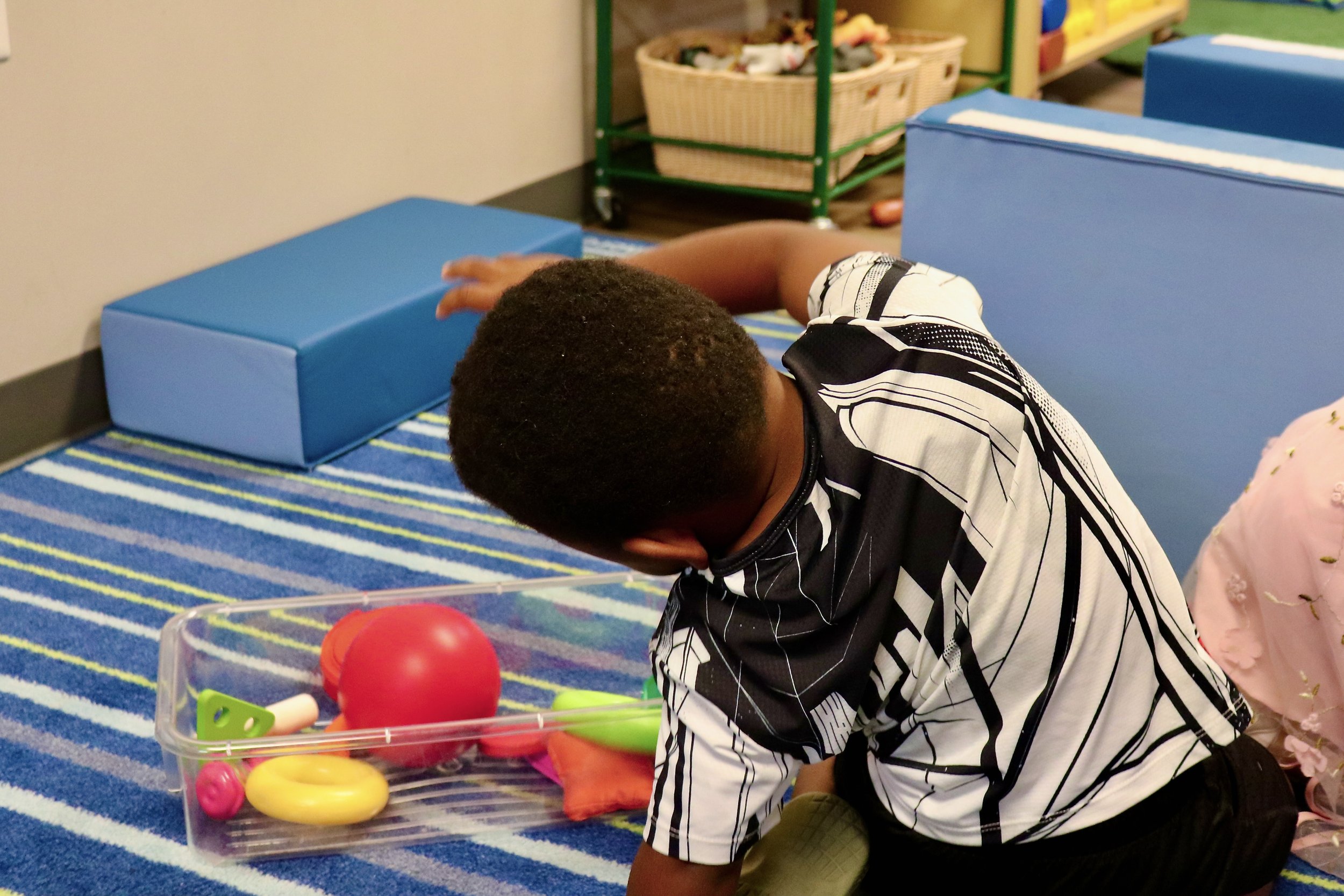A young boy with short curly hair kneeling on a colorful striped rug, reaching for toys in a clear plastic container, with blue padded blocks and a basket of toys in the background.