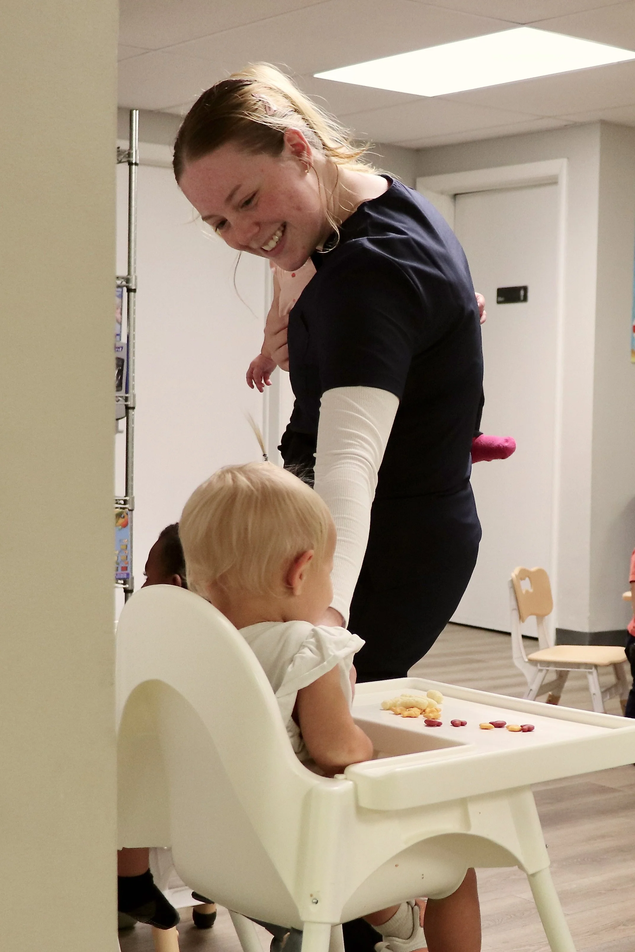 A woman with long hair, smiling, leaning over a child sitting in a high chair in a room with light-colored walls and ceiling panels. The child is playing with colorful candy on a tray. In the background, there are small chairs and a door.
