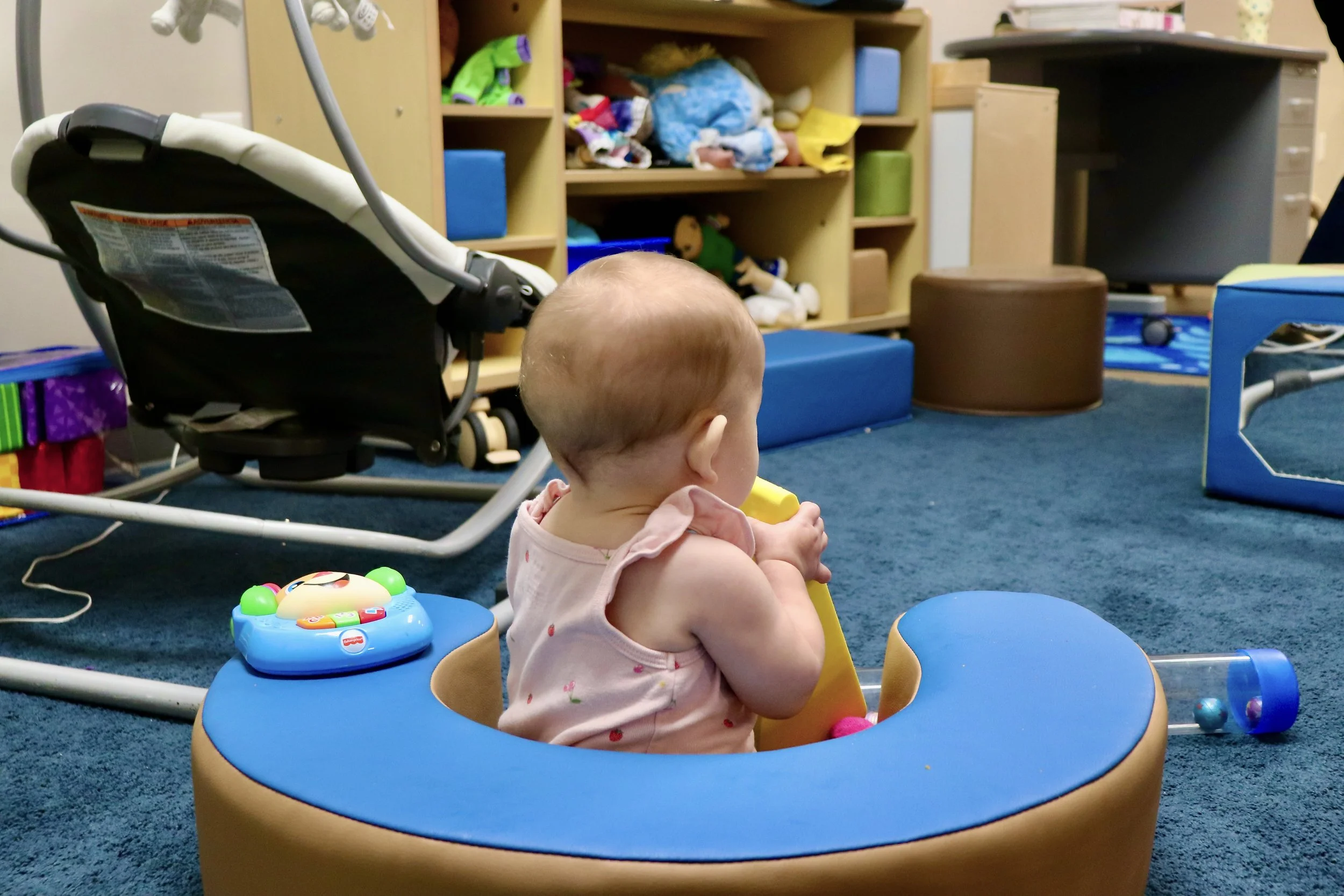 A baby sitting in a blue and yellow activity center in a playroom with shelves filled with toys, blankets, and other items. The baby is holding a yellow toy and has a pacifier in the activity center.
