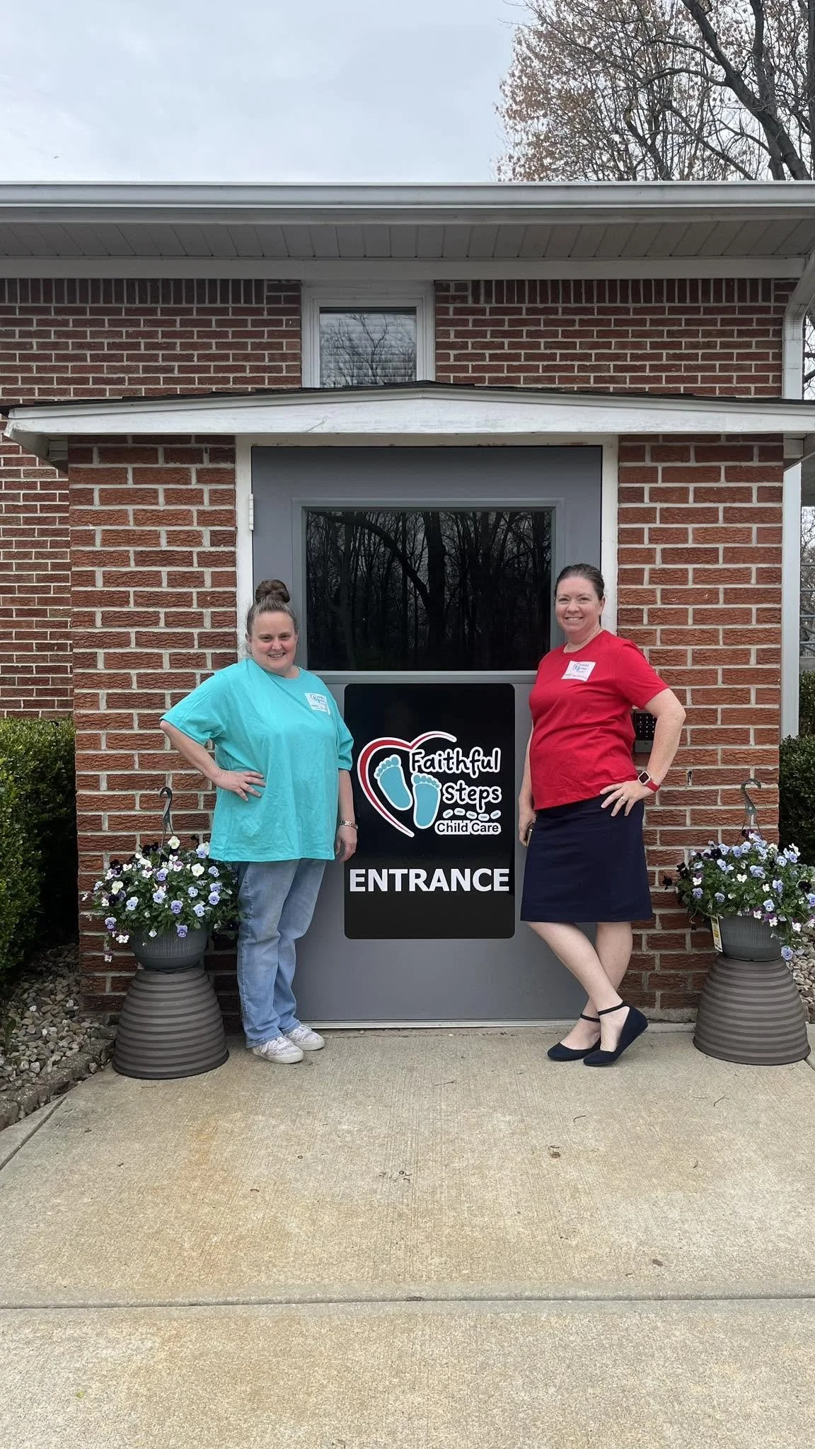 Two women standing outside in front of a sign that reads 'Faithful Steps Child Care' at the entrance of a brick building. The woman on the left is wearing a light blue shirt and jeans, while the woman on the right is wearing a red top, black skirt, and black shoes. There are two flower pots with purple and white flowers on either side of the sign.