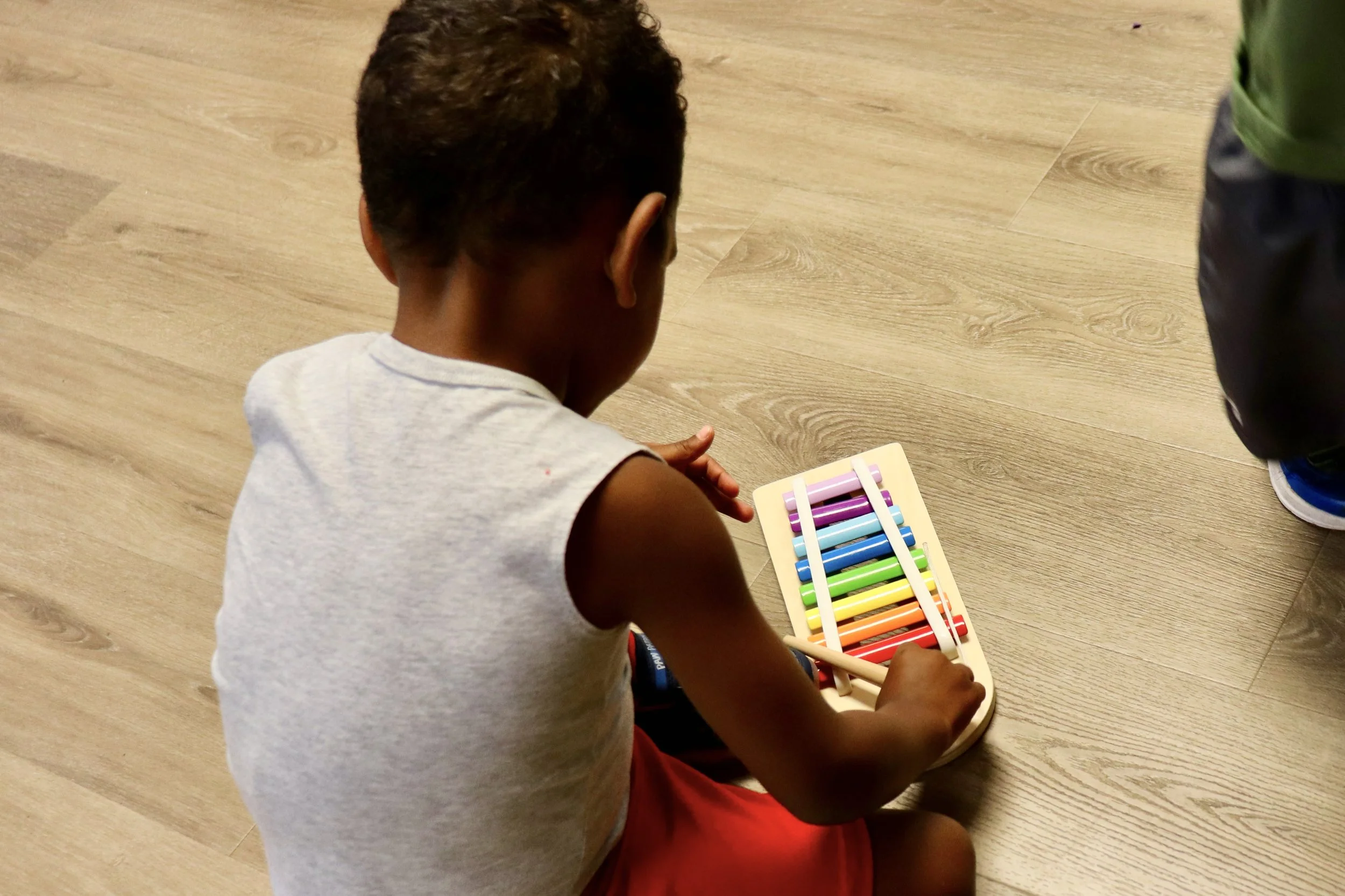 A child sitting on the floor playing with a rainbow-colored xylophone.