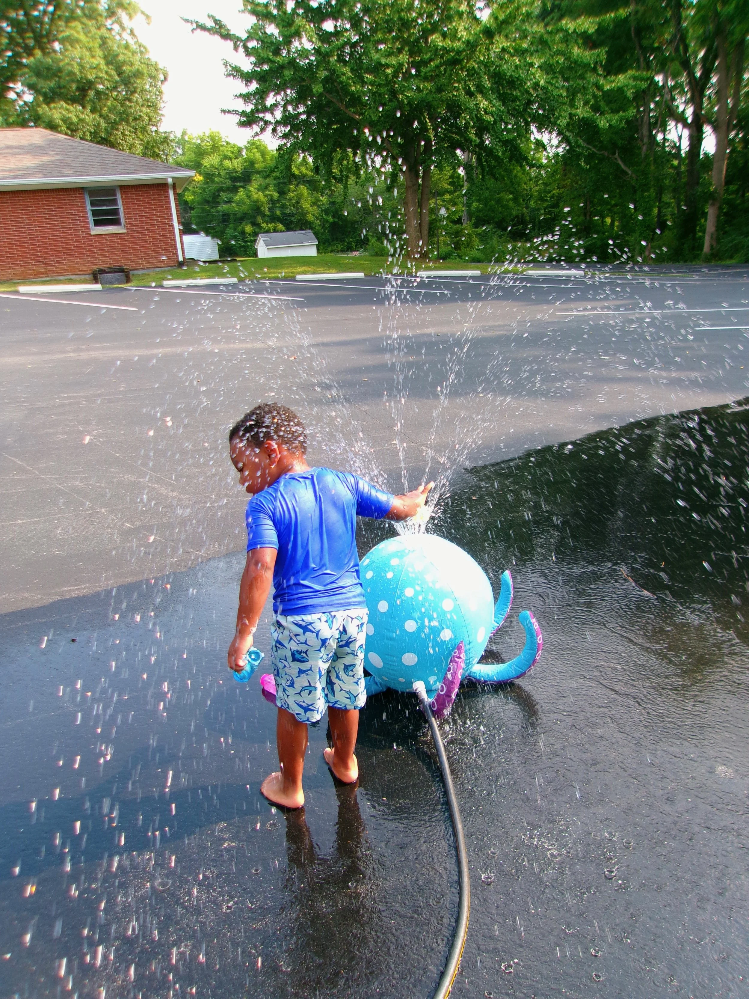 A young boy wearing a blue swim shirt and patterned swim shorts is playing in a puddle with a large blue octopus-shaped water toy. He is holding a garden hose, spraying water, causing a splash on the wet pavement. In the background, there are trees, a brick building, and a white shed.